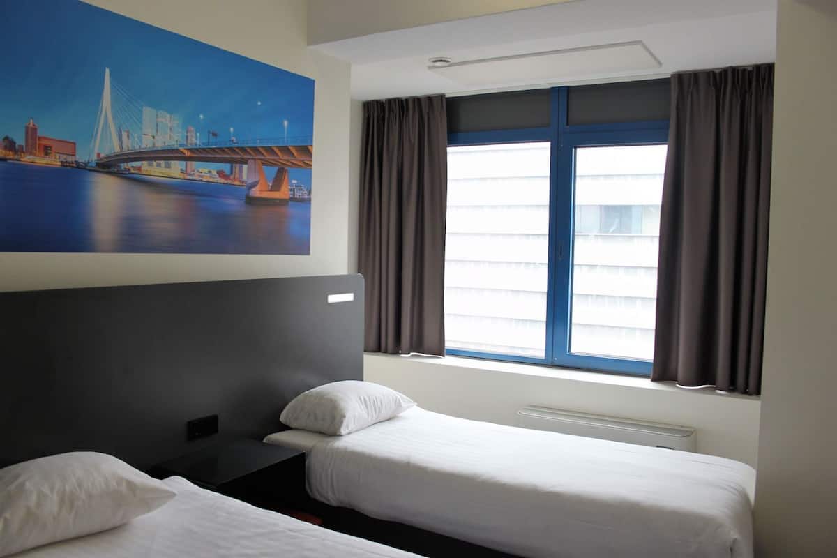 A hotel room with two single beds, a large window with curtains, a painting of a bridge, and a white ceiling.