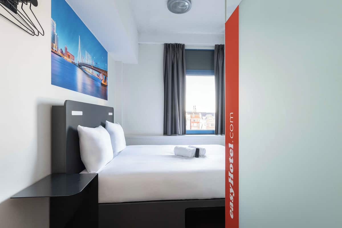 A hotel room with a bed, a window with curtains, a painting on the wall, and a red sign with text.