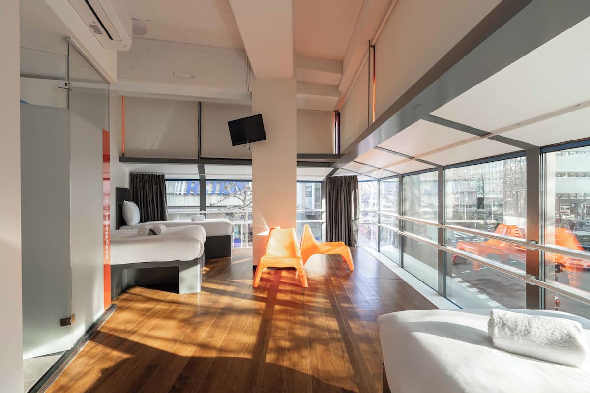 A modern hotel room with a large window, a sofa, a small table, and a TV mounted on the wall.