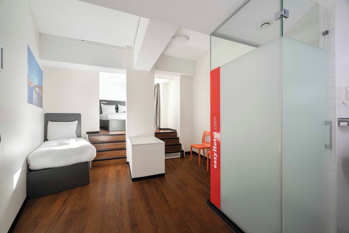 A modern hotel room with a bed, a small seating area, a staircase, and a glass partition.
