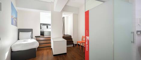 A modern hotel room with a bed, a small seating area, a staircase, and a glass partition.