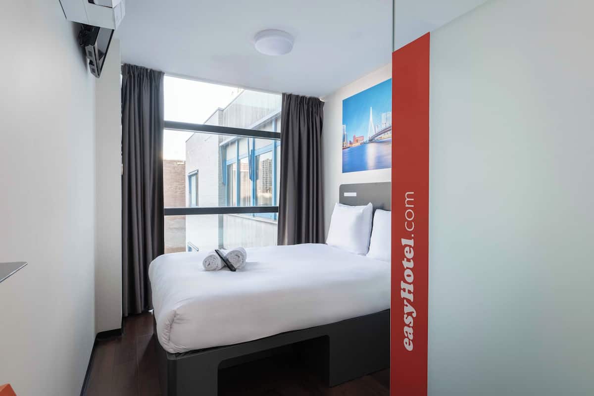 A hotel room with a bed, a window with curtains, a wall-mounted air conditioning unit, and a painting of a bridge.