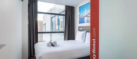 A hotel room with a bed, a window with curtains, a wall-mounted air conditioning unit, and a painting of a bridge.