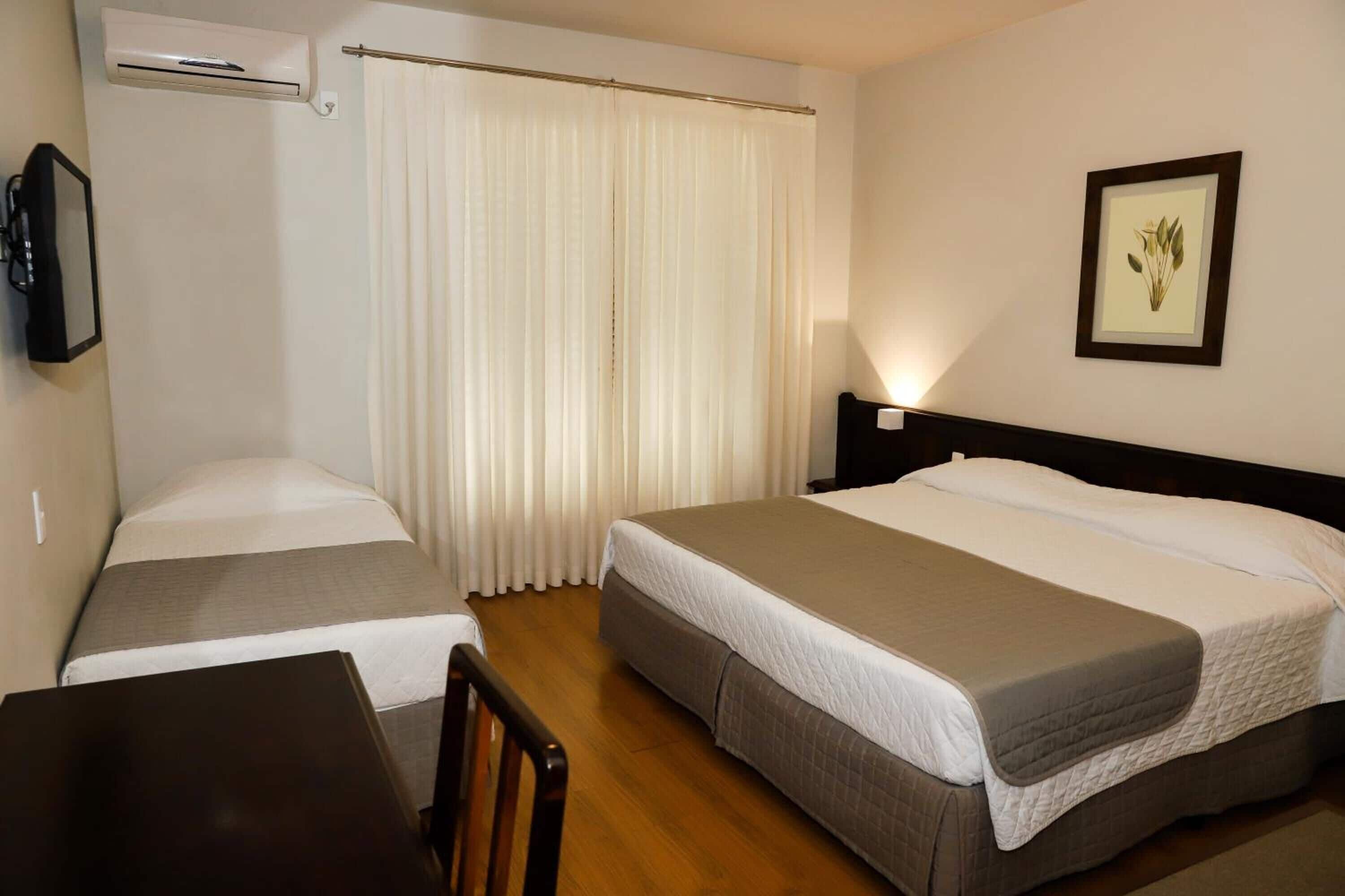 Habitación de hotel con dos camas, un escritorio, una silla, aire acondicionado, cortina, un cuadro en la pared y una lámpara de noche.