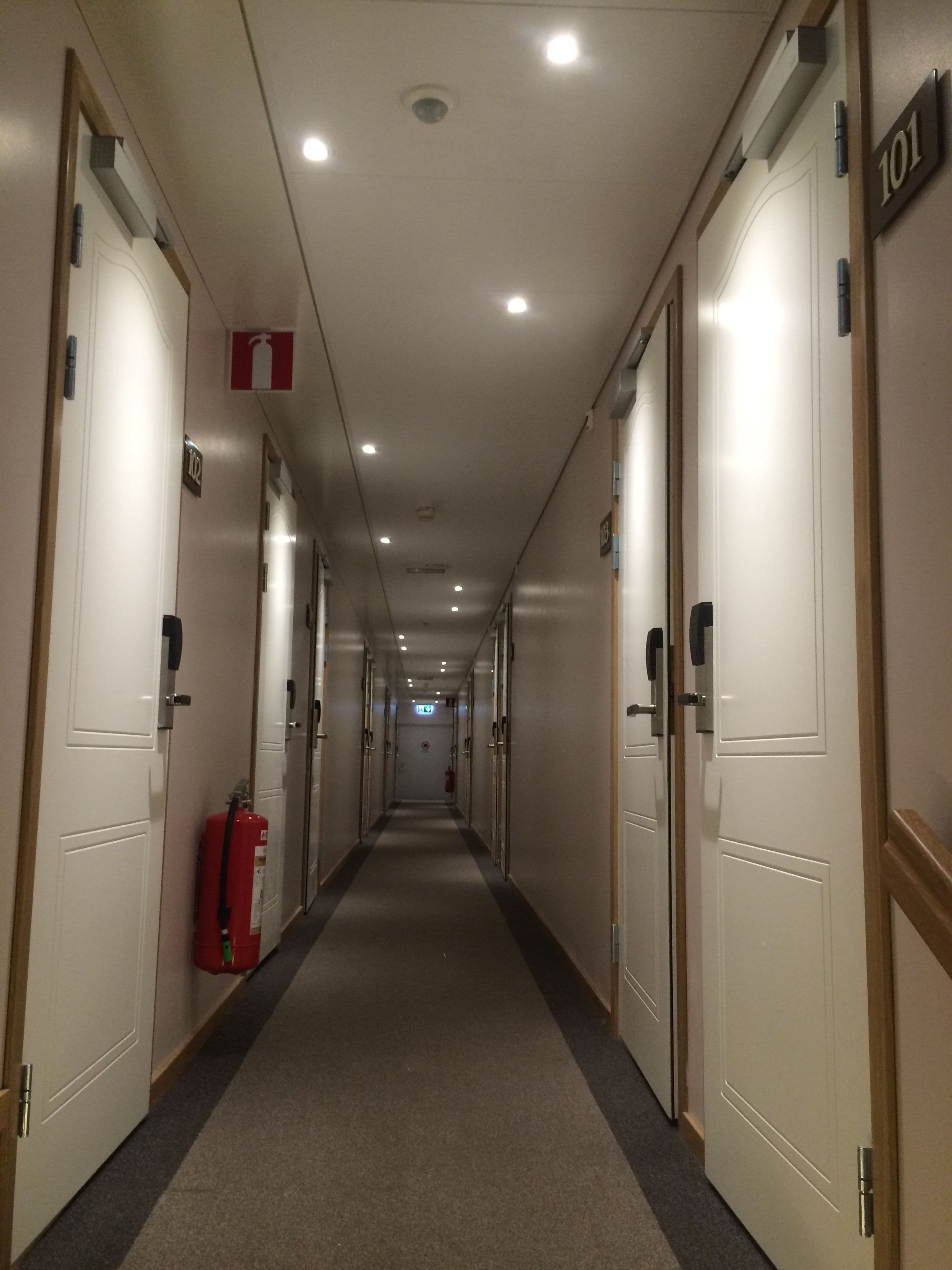 Un pasillo largo y estrecho de hotel con puertas a ambos lados, con los números 101 y 102, y un extintor en la pared izquierda.