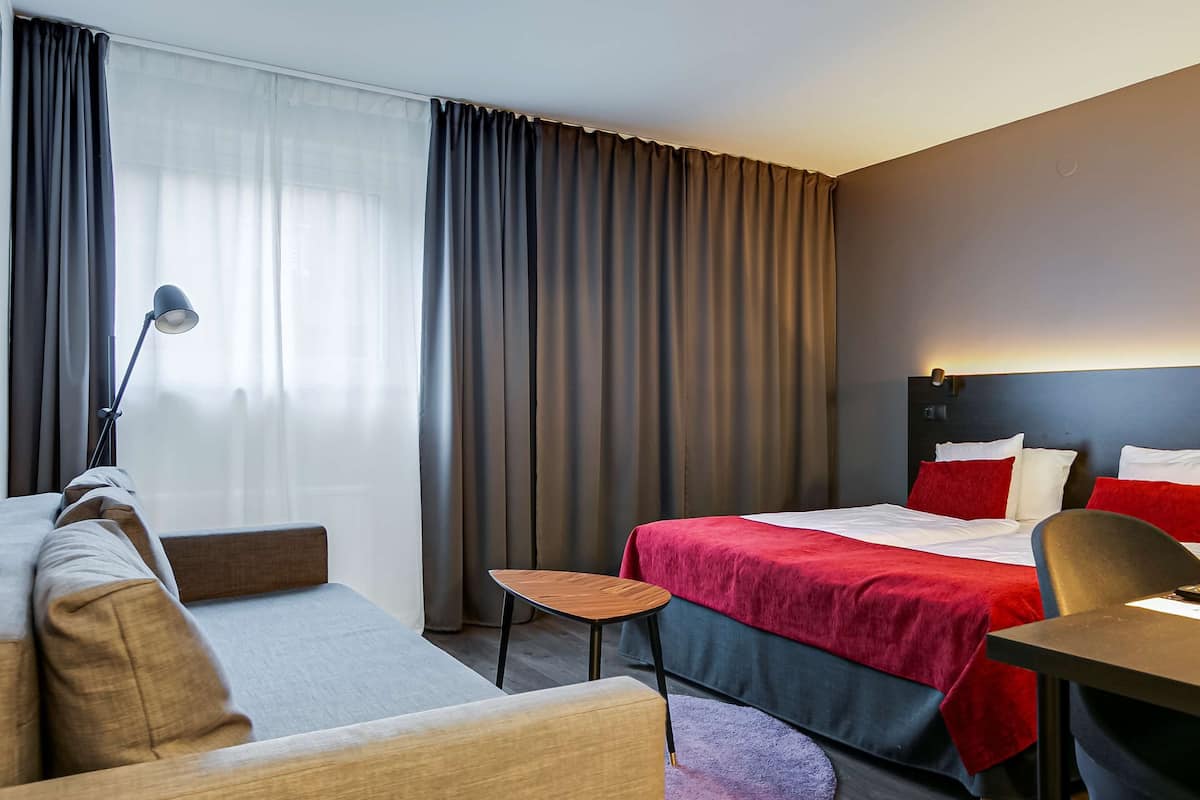 A hotel room with a bed, a desk, a chair, a small table, and a lamp.