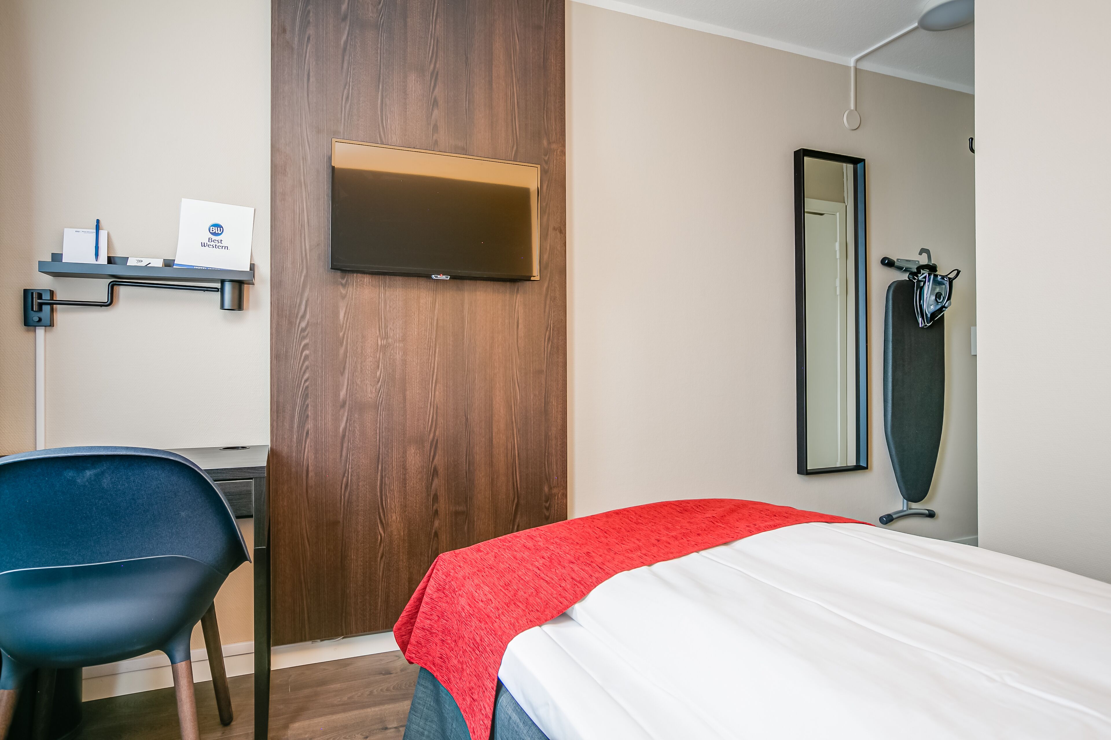 Een hotelkamer met een bed, een bureau, een stoel, een televisie, een spiegel en een handdoekrek.