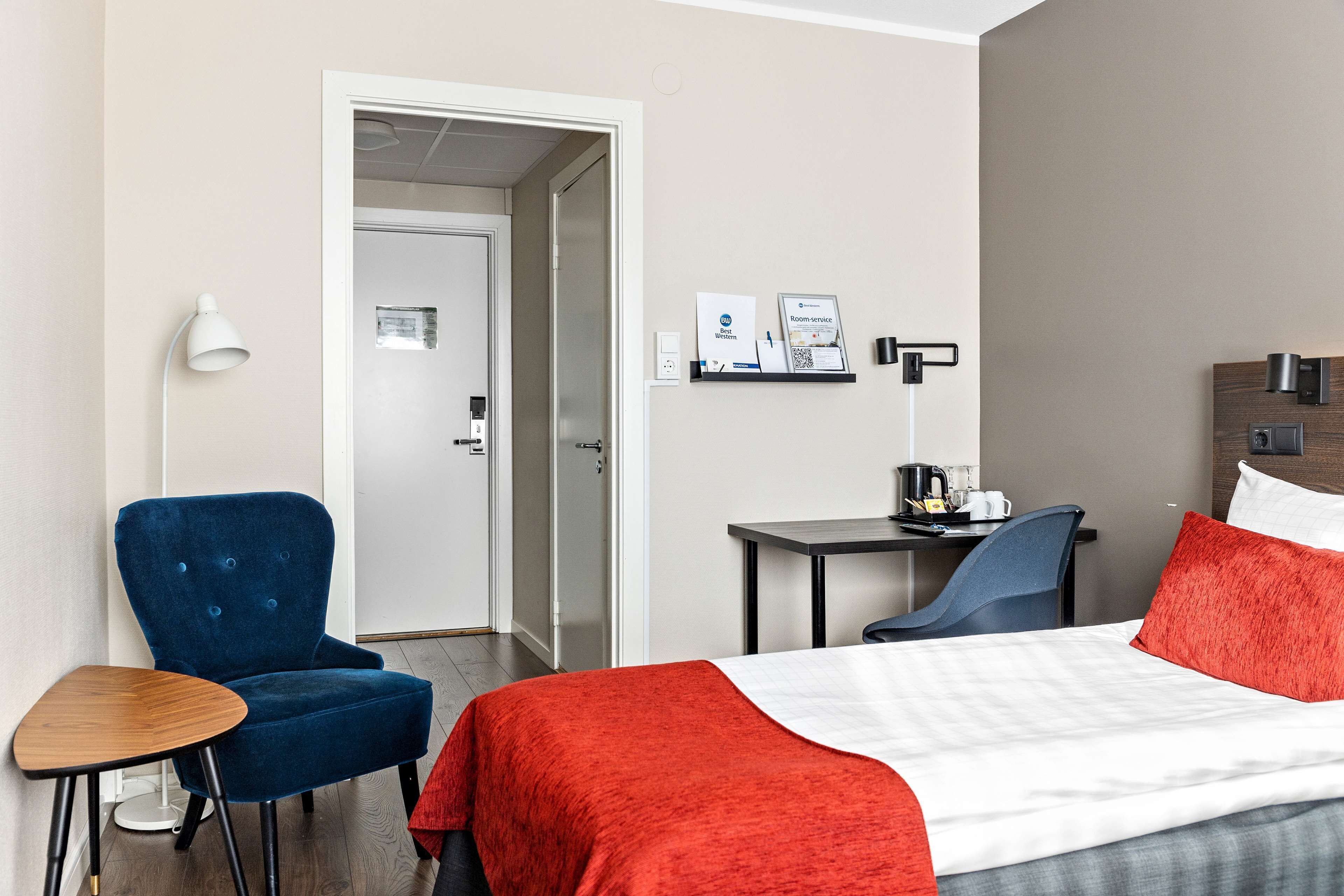 A hotel room with a bed, a blue armchair, a small table, a desk, and a coffee machine.