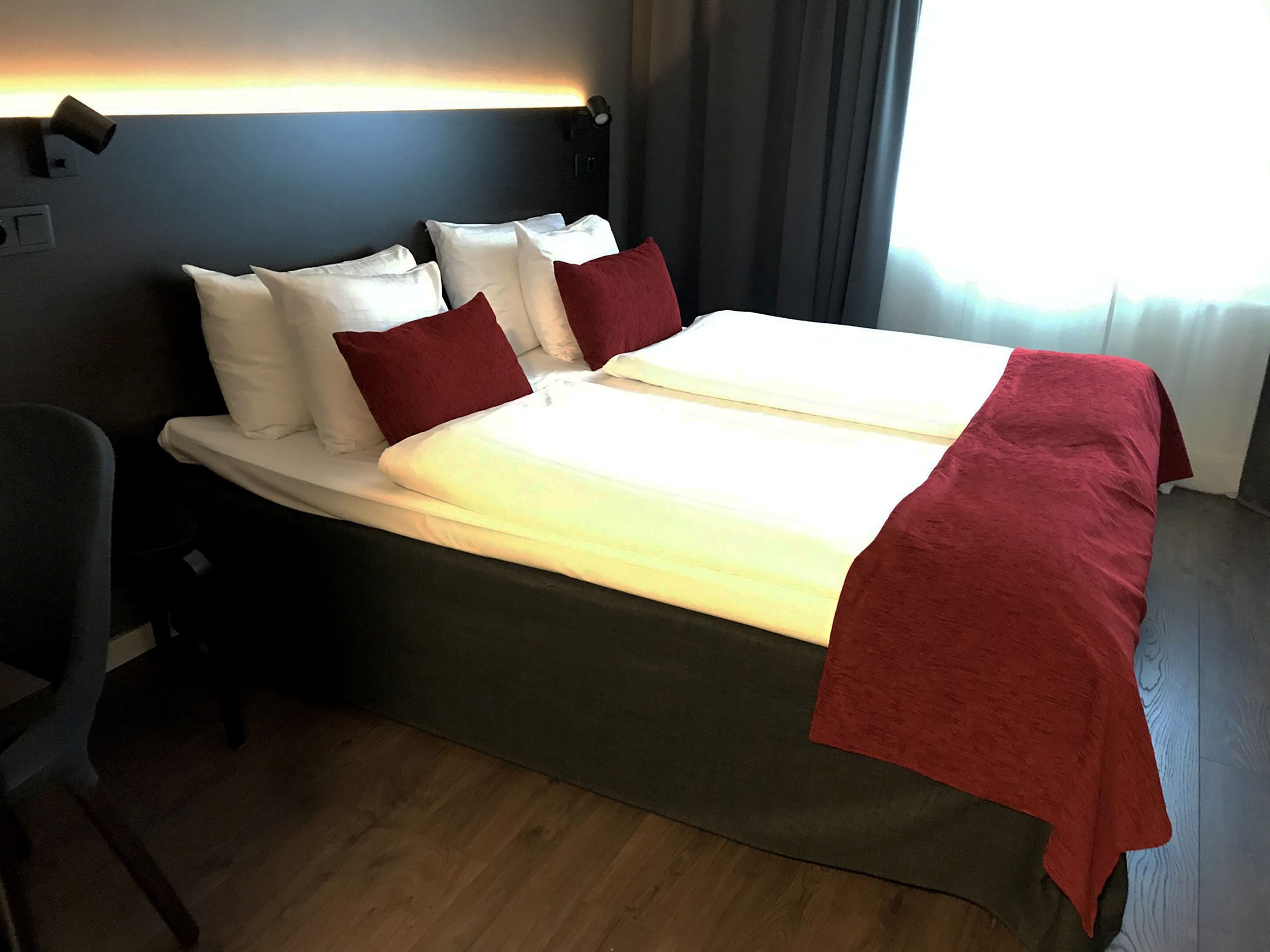 A hotel room with a large bed, two white pillows, a red throw blanket, and a headboard with a light fixture.