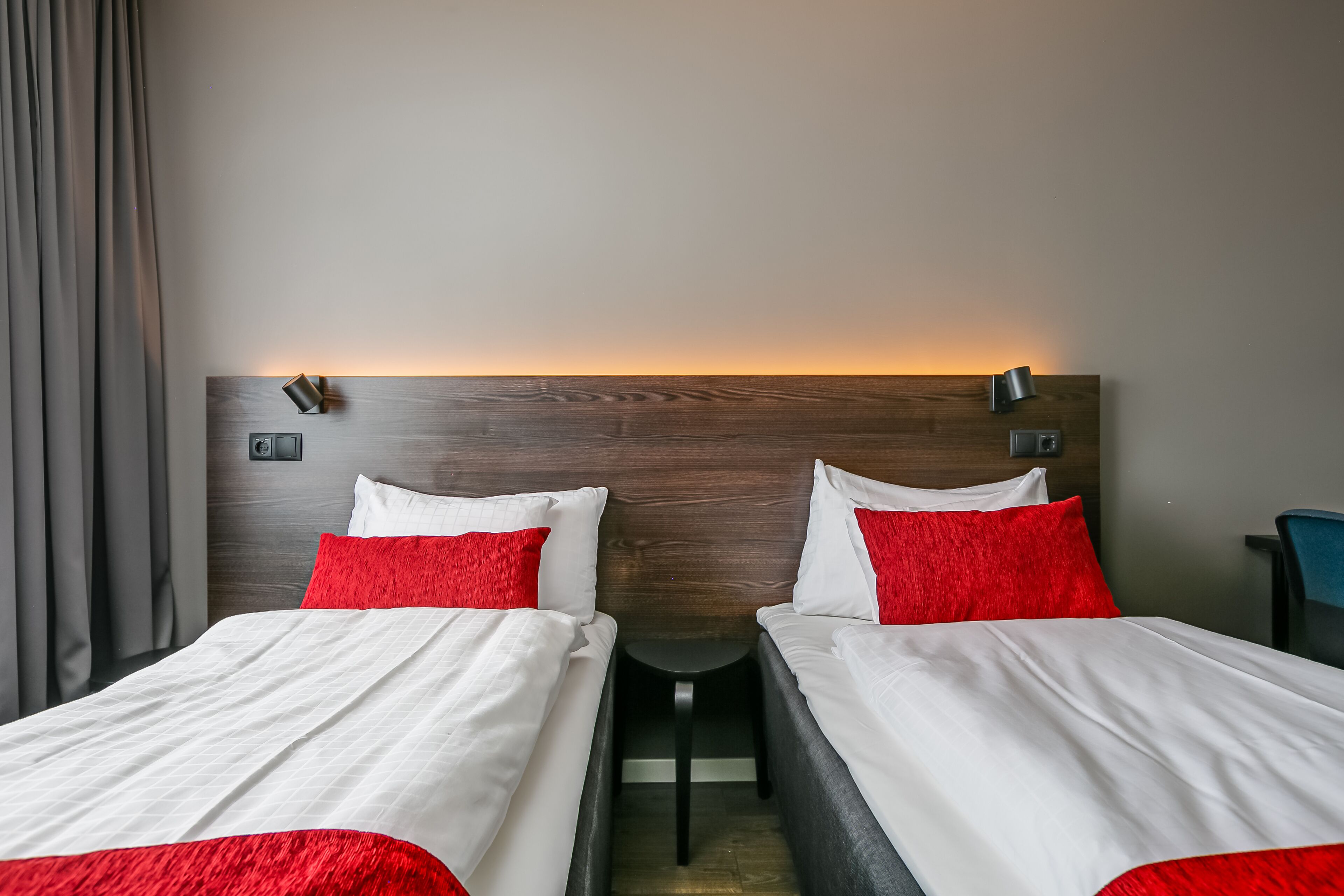 Une chambre d’hôtel avec deux lits, chacun agrémenté de draps blancs et d’oreillers rouges, d’une tête de lit en bois et d’une petite table placée entre les lits.