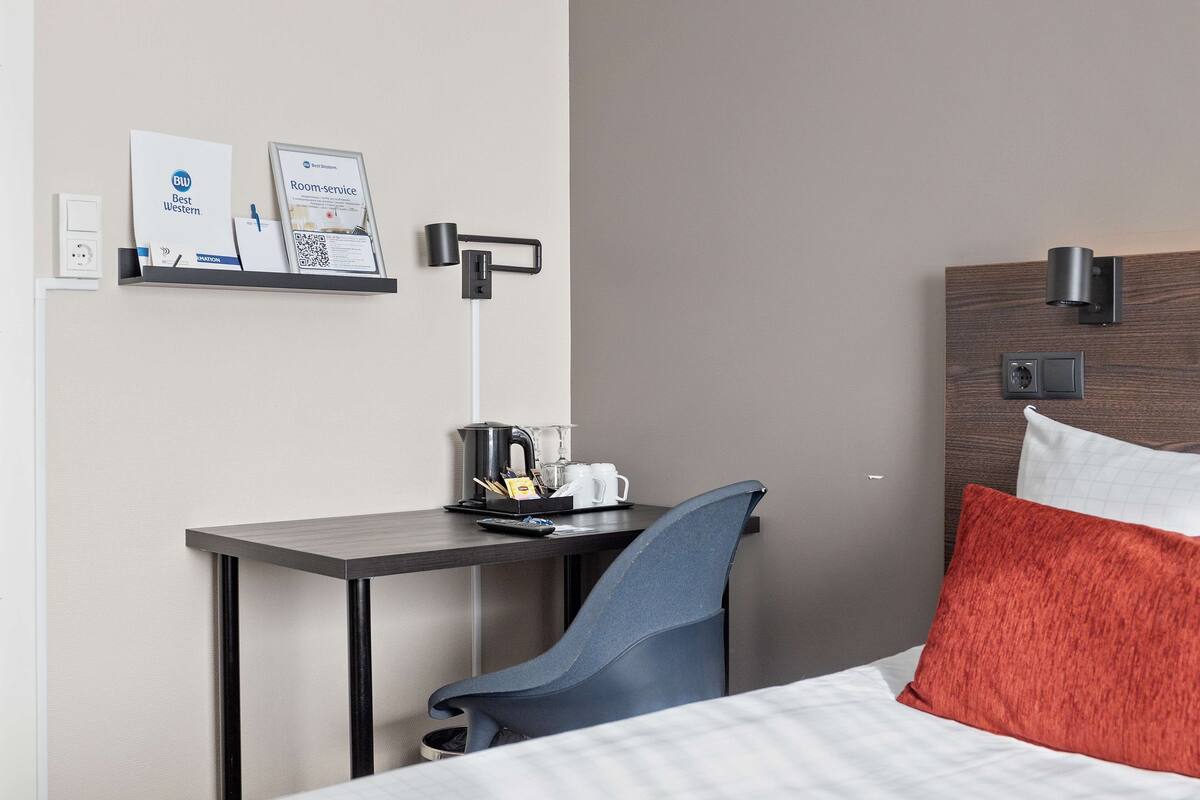 A hotel room with a bed, a desk with a chair, a coffee maker, and a shelf with informational materials.