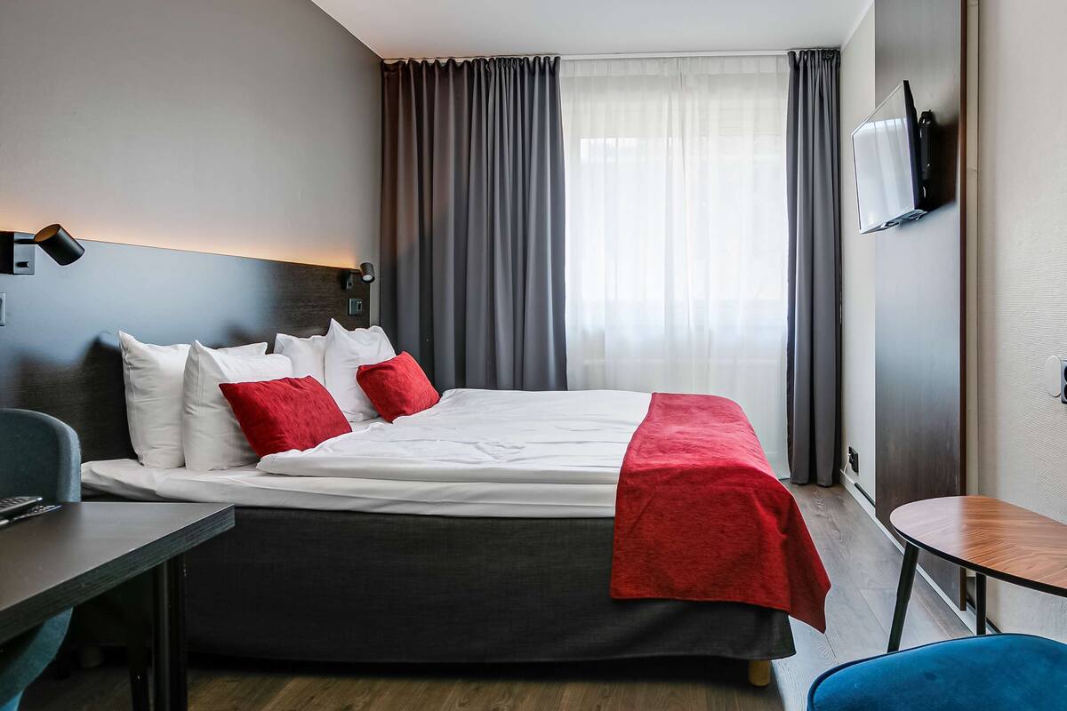 A modern hotel room with a large bed, a desk, a chair, and a TV mounted on the wall.