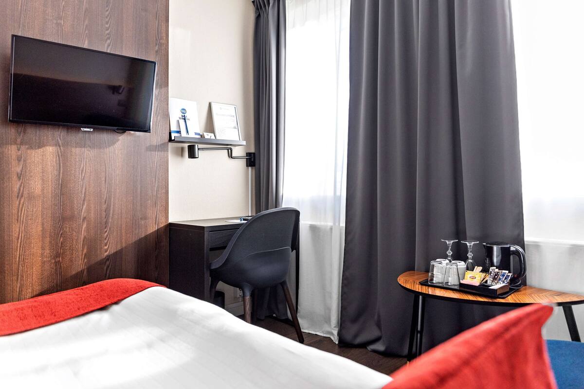 A hotel room with a bed, a desk, a chair, a TV mounted on the wall, and a small table with a tray of glasses and a coffee pot.