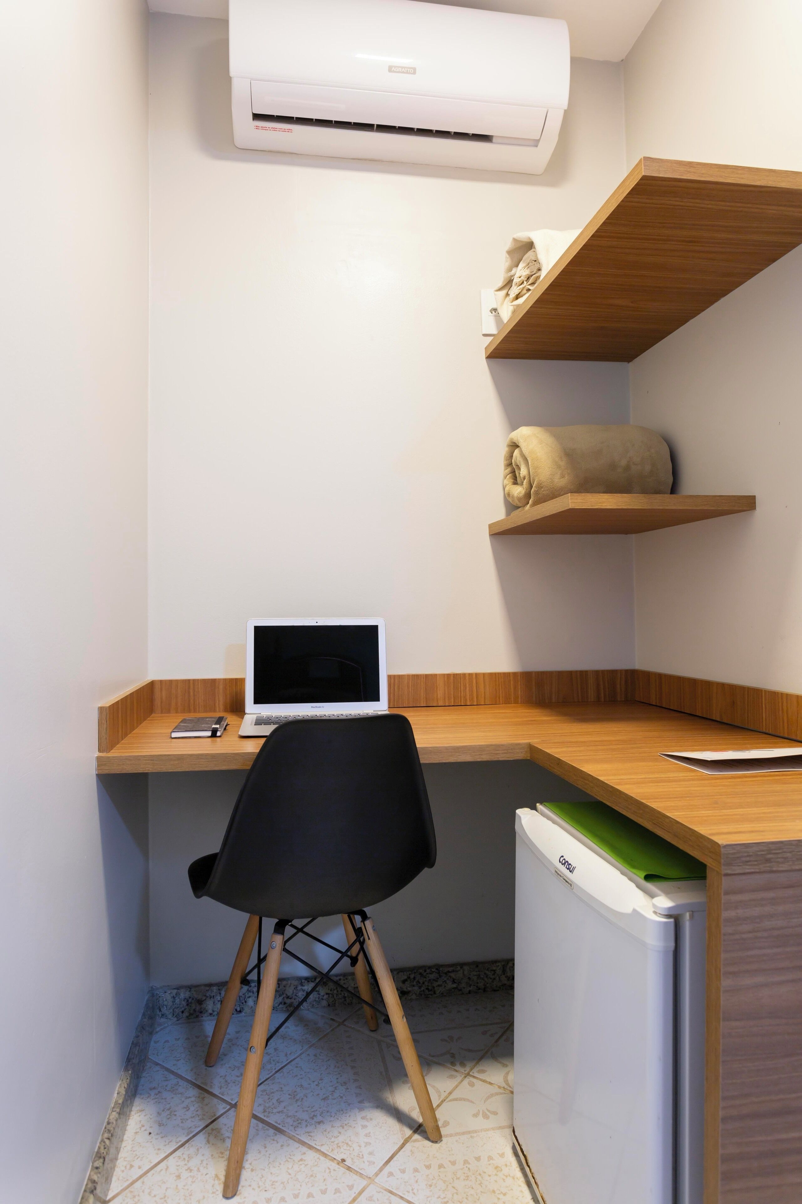 A small, modern kitchenette with a white refrigerator, a laptop on a wooden desk, and a black chair.