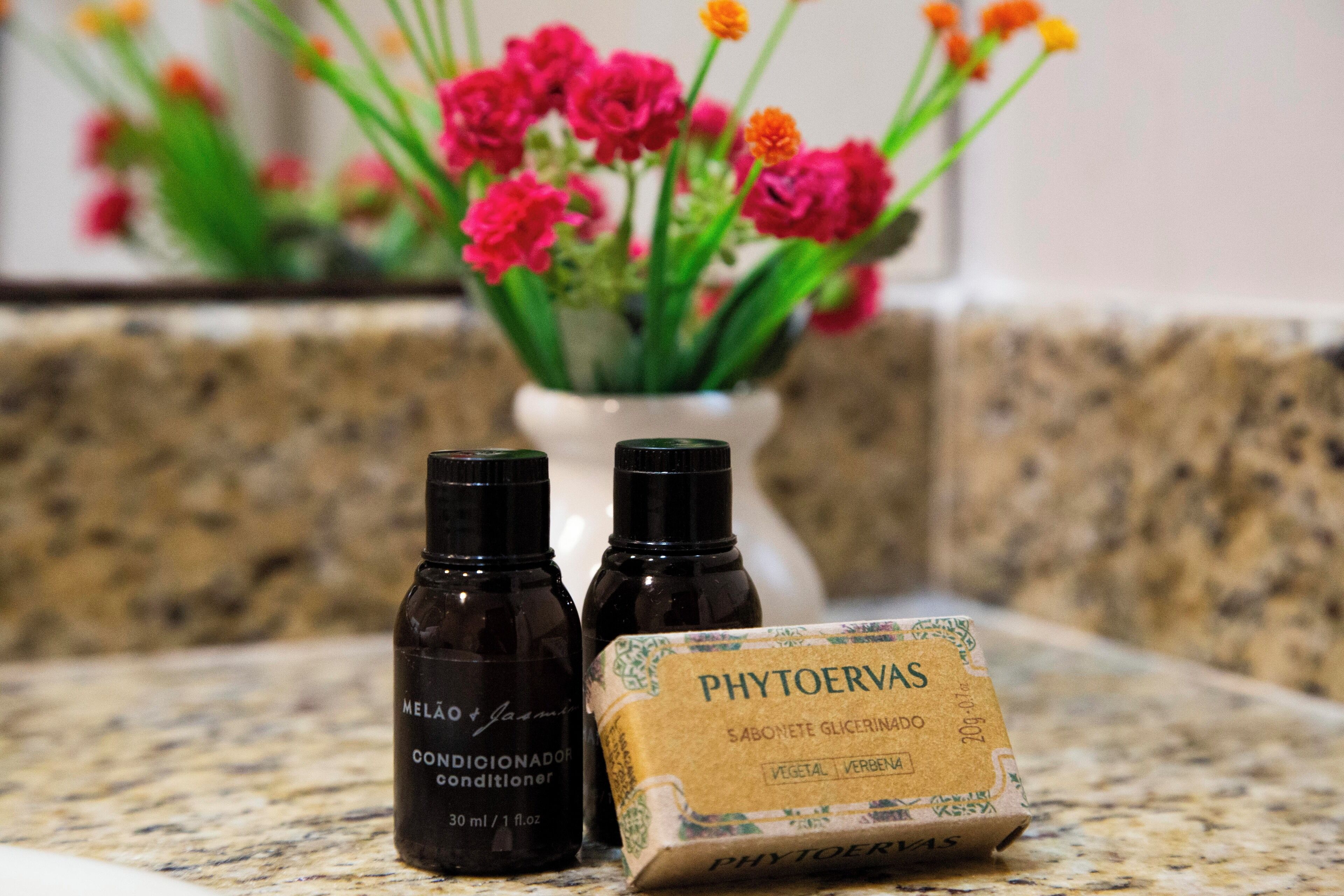 Een aanrecht met twee donkerbruine flessen met de tekst 'MELAO & Jasmine Conditioner' en een doosje met de tekst 'PHYTOERVAS'. In de achtergrond staat een vaas met roze en oranje bloemen.