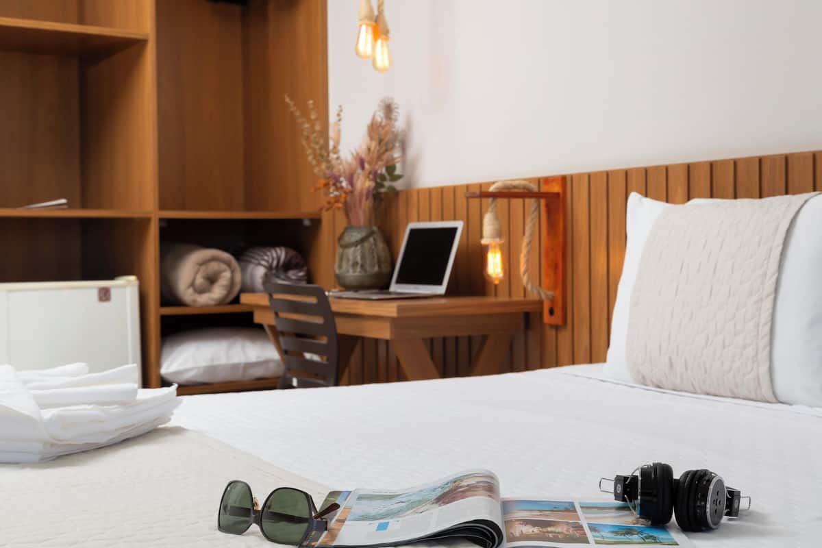 A neatly made bed with a magazine, sunglasses, and headphones on it. There is a wooden desk with a laptop and a vase with dried flowers.