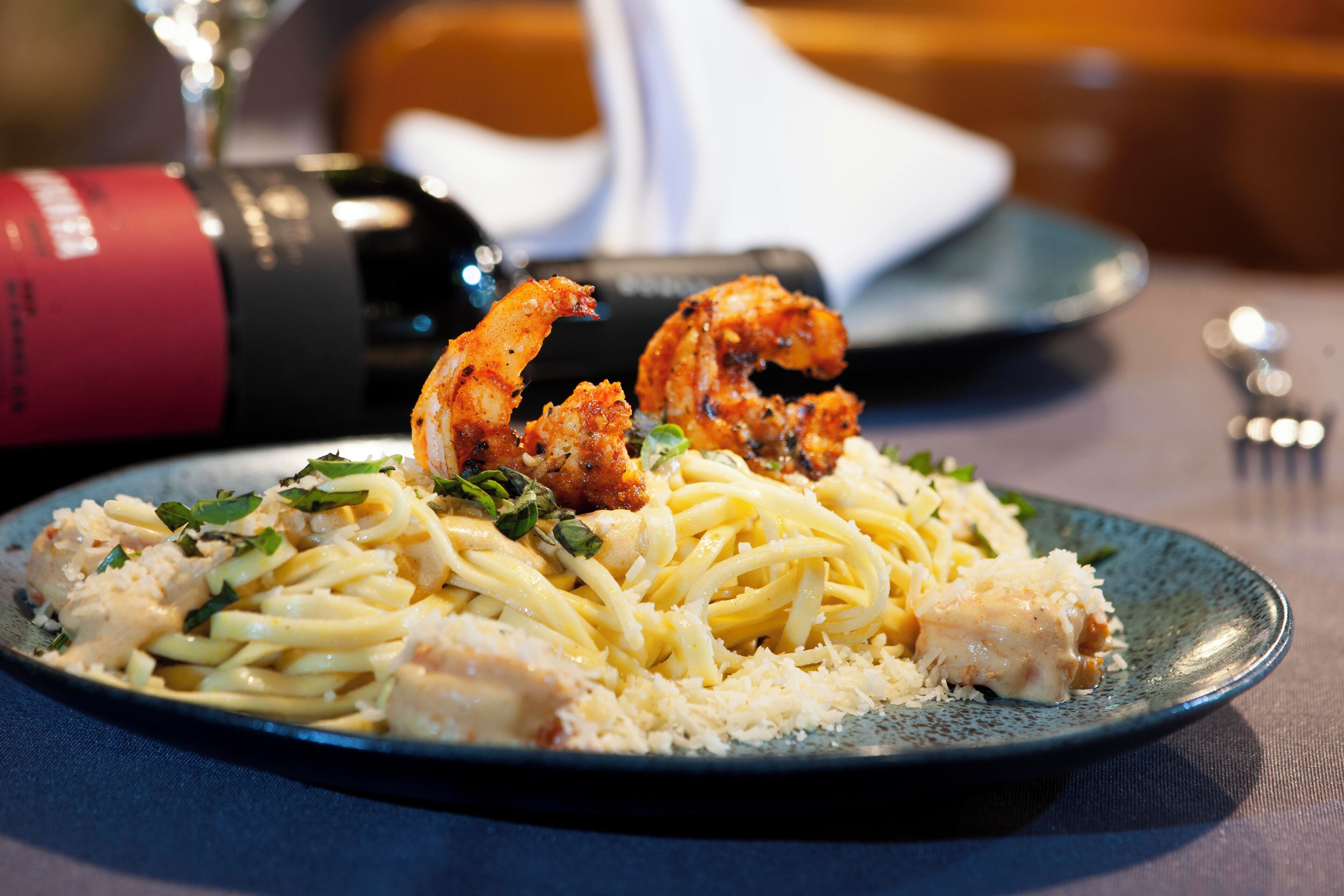 A plate of pasta with shrimp and chicken, a wine bottle, and a fork on a table.