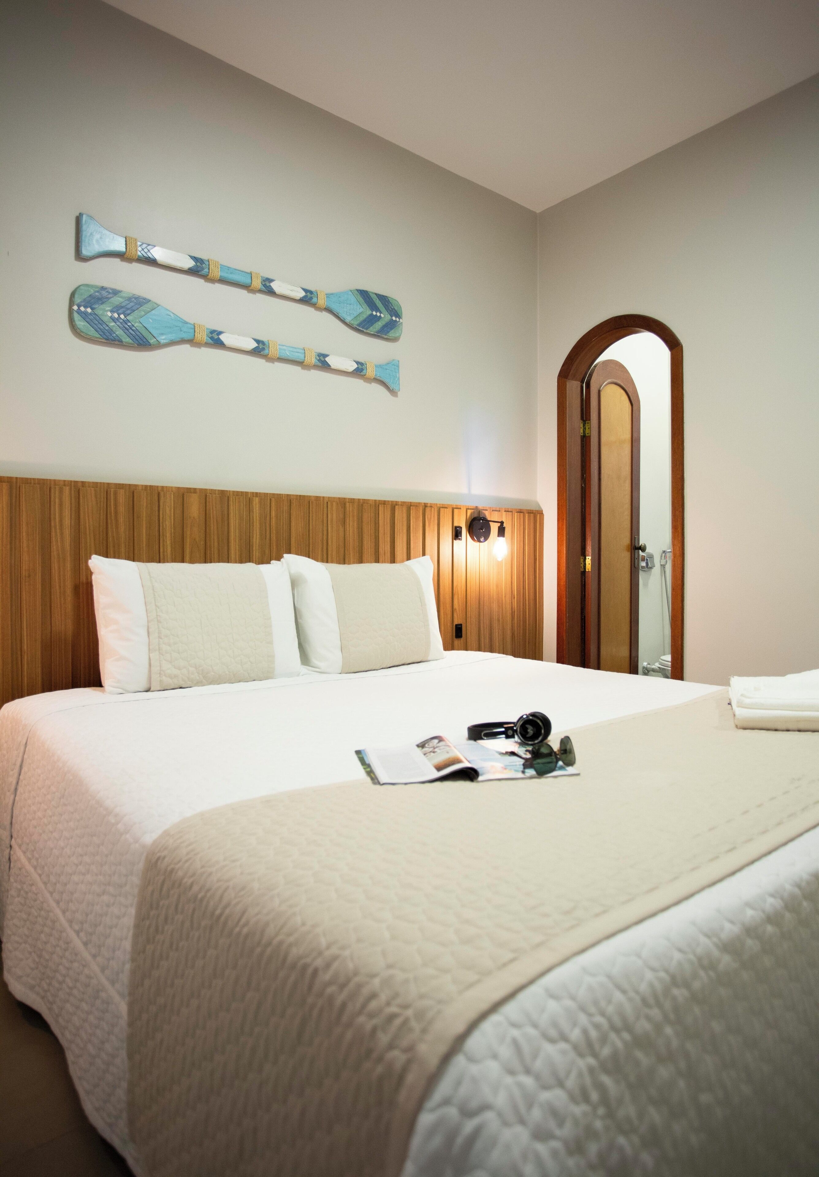 A hotel room with a wooden headboard, a large bed with white bedding, a mirror, and a pair of decorative paddles on the wall.