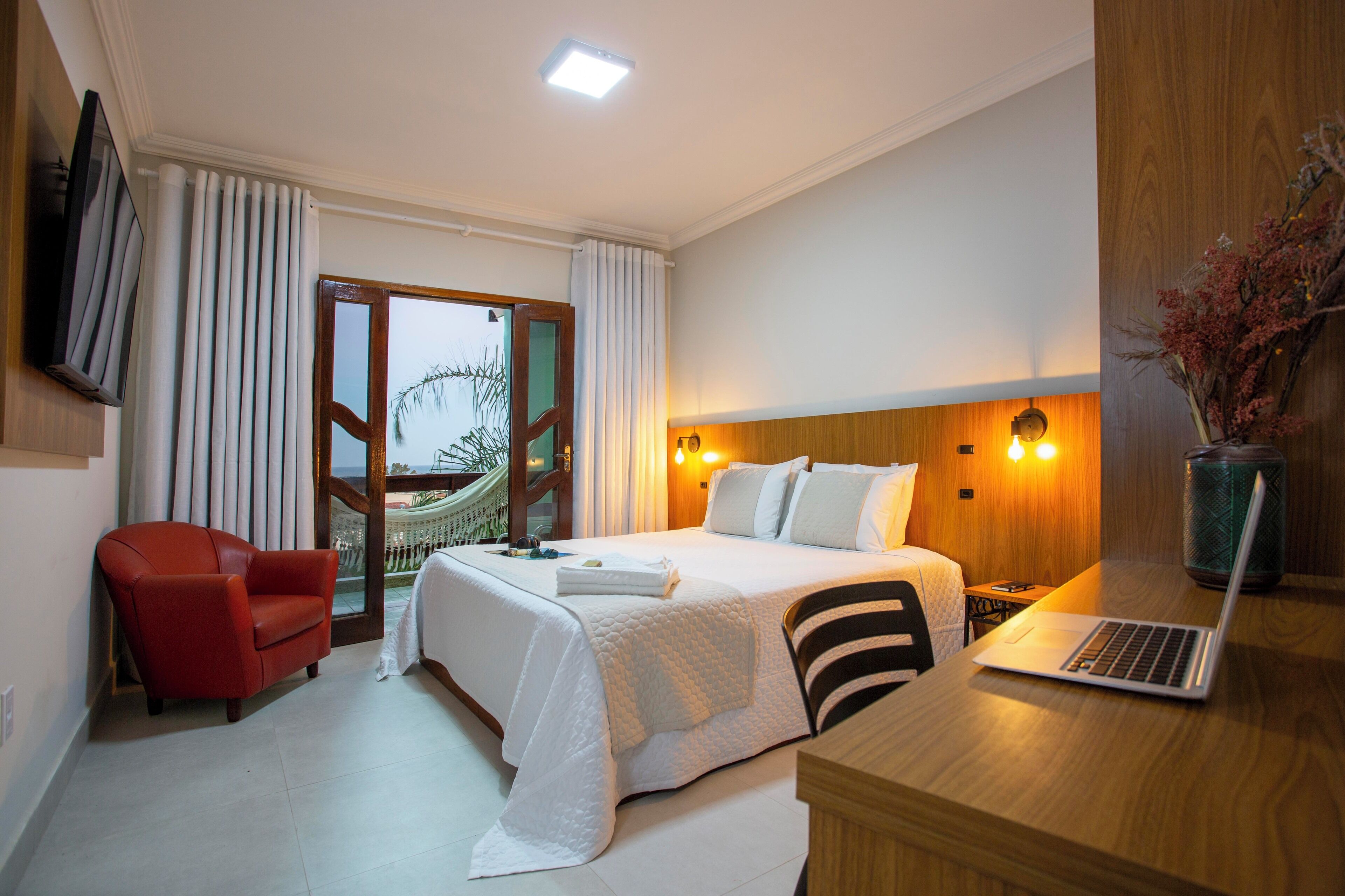 A hotel room with a bed, a desk with a laptop, a red chair, and a view of a building outside.
