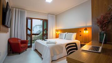 A hotel room with a bed, a desk with a laptop, a red chair, and a view of a building outside.