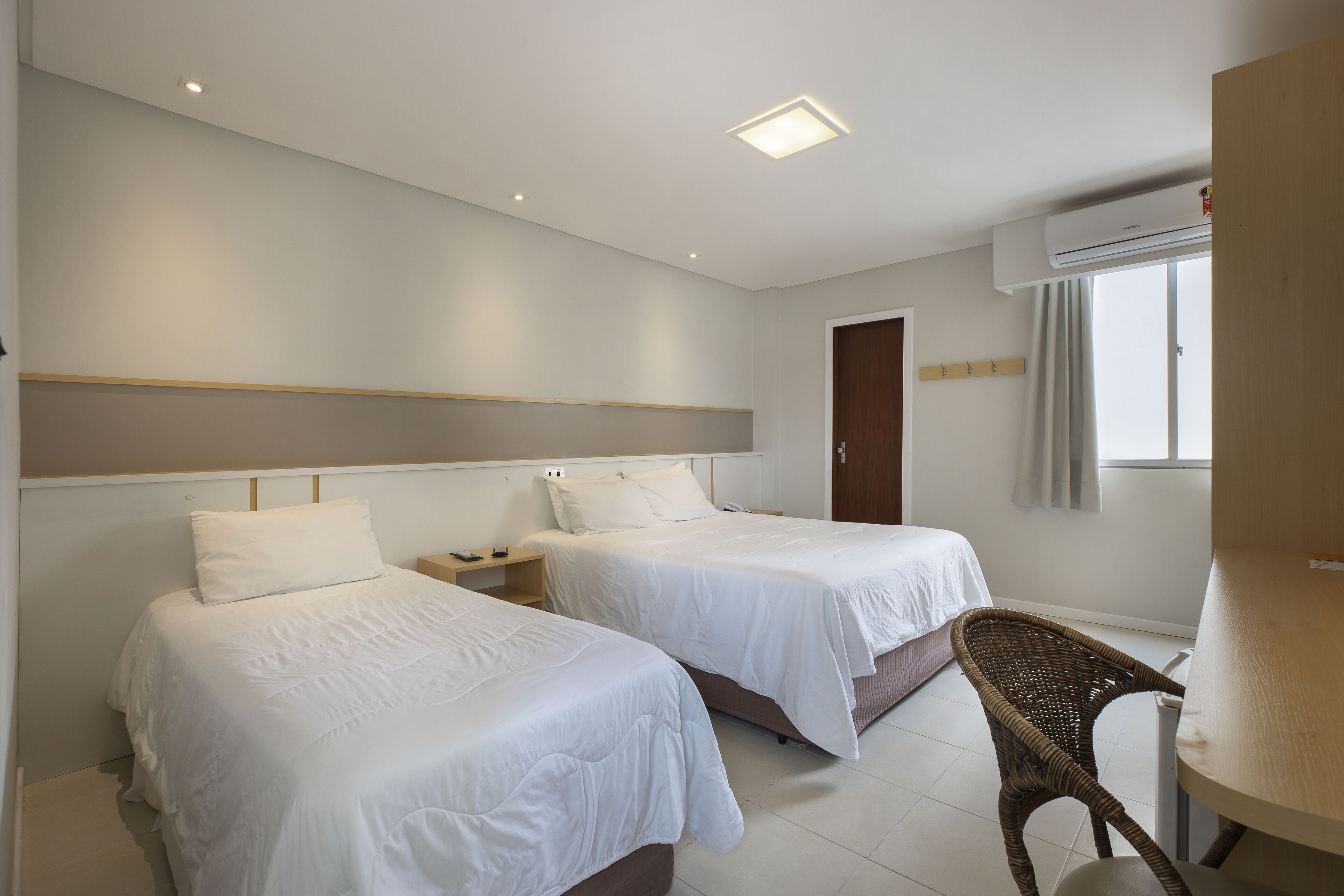 A hotel room with two beds, a small table, a chair, an air conditioning unit, and a window with curtains.