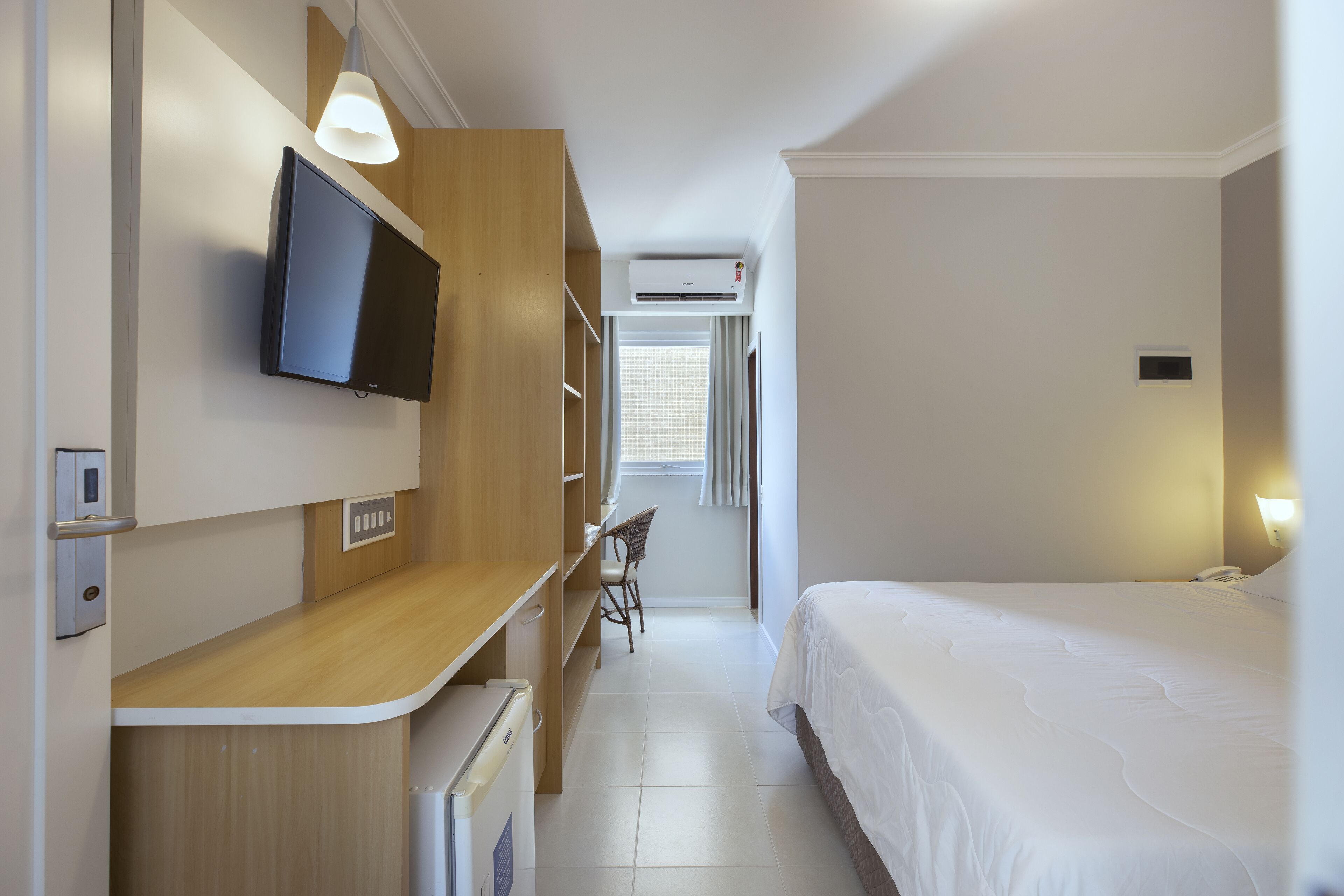 A hotel room with a bed, a desk with a chair, a TV, an air conditioning unit, and a window with curtains.
