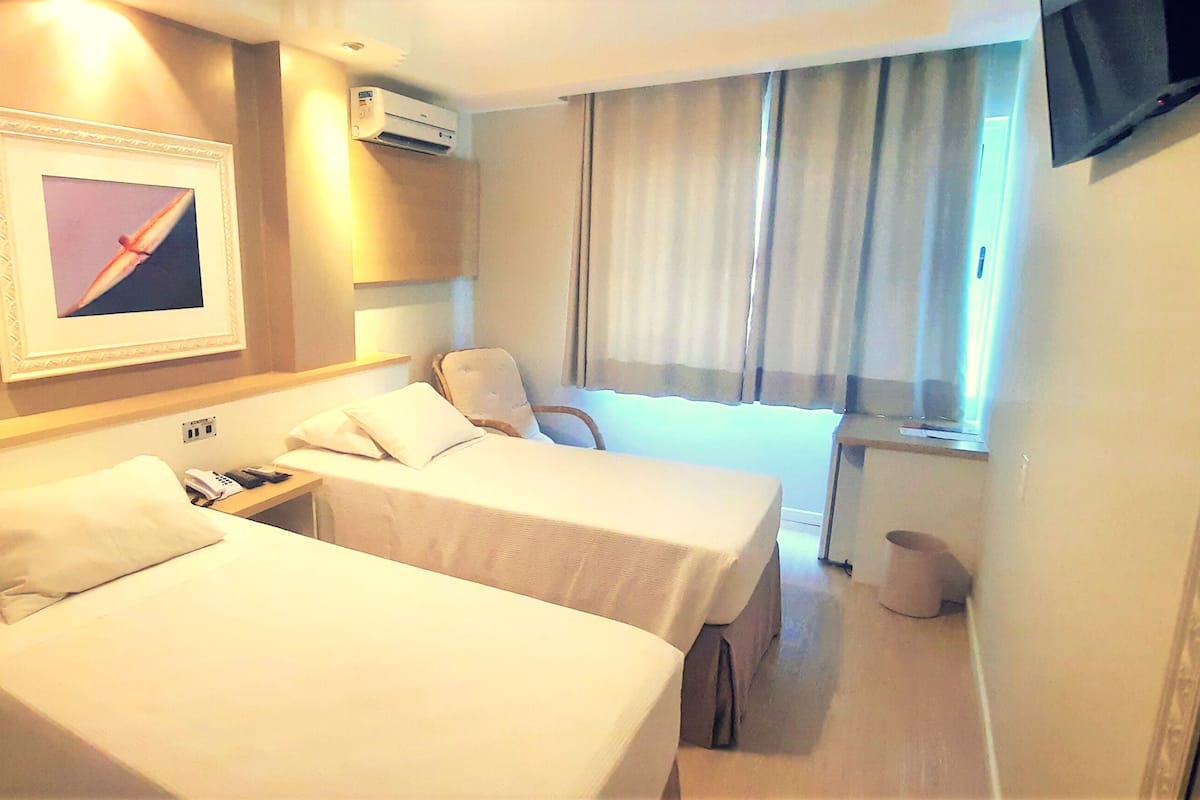 A hotel room with two beds, a TV, an air conditioning unit, and a window with curtains.