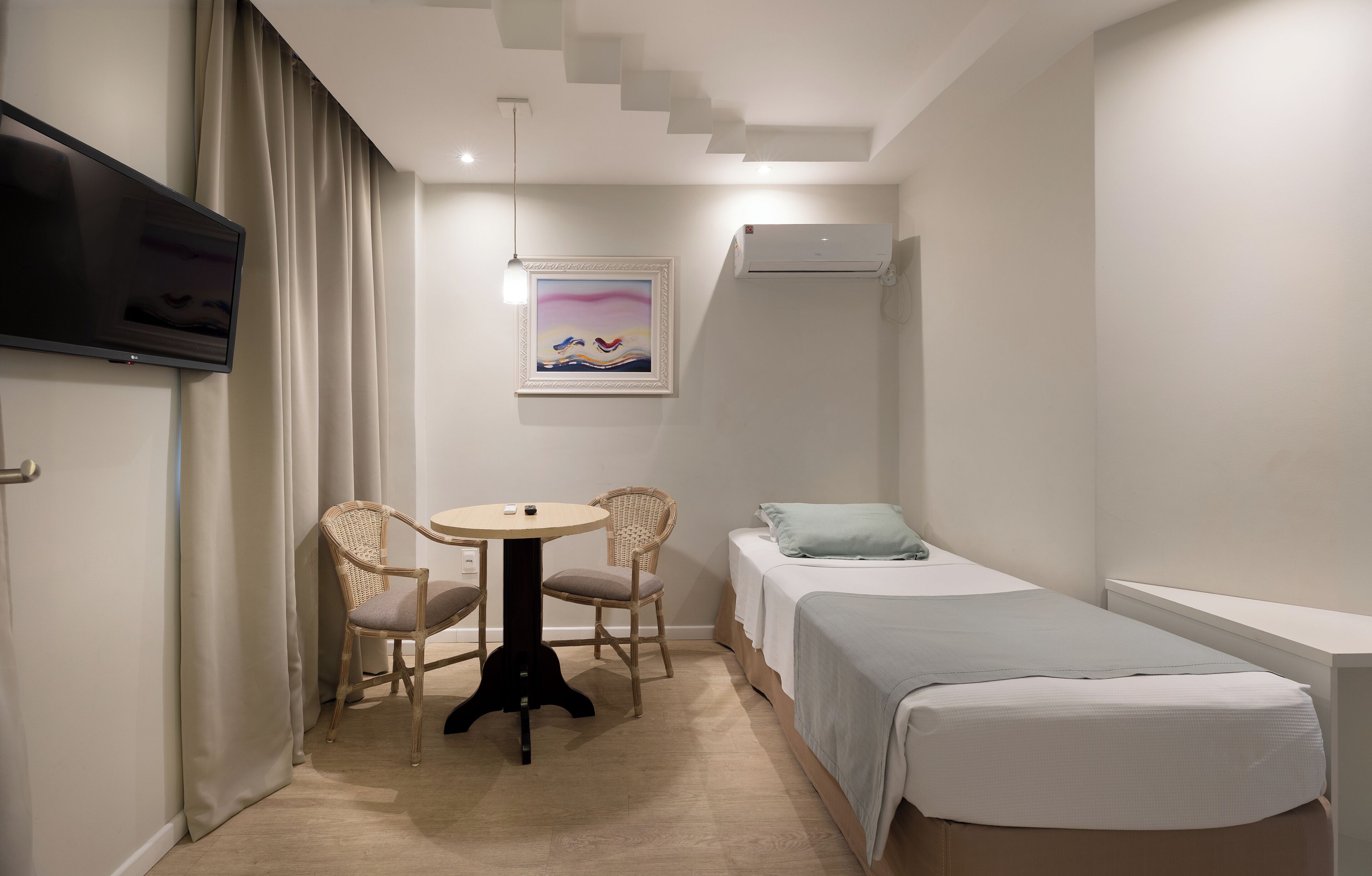 Una camera d'albergo compatta con un letto, un tavolino, due sedie, una televisione e un quadro appeso al muro.