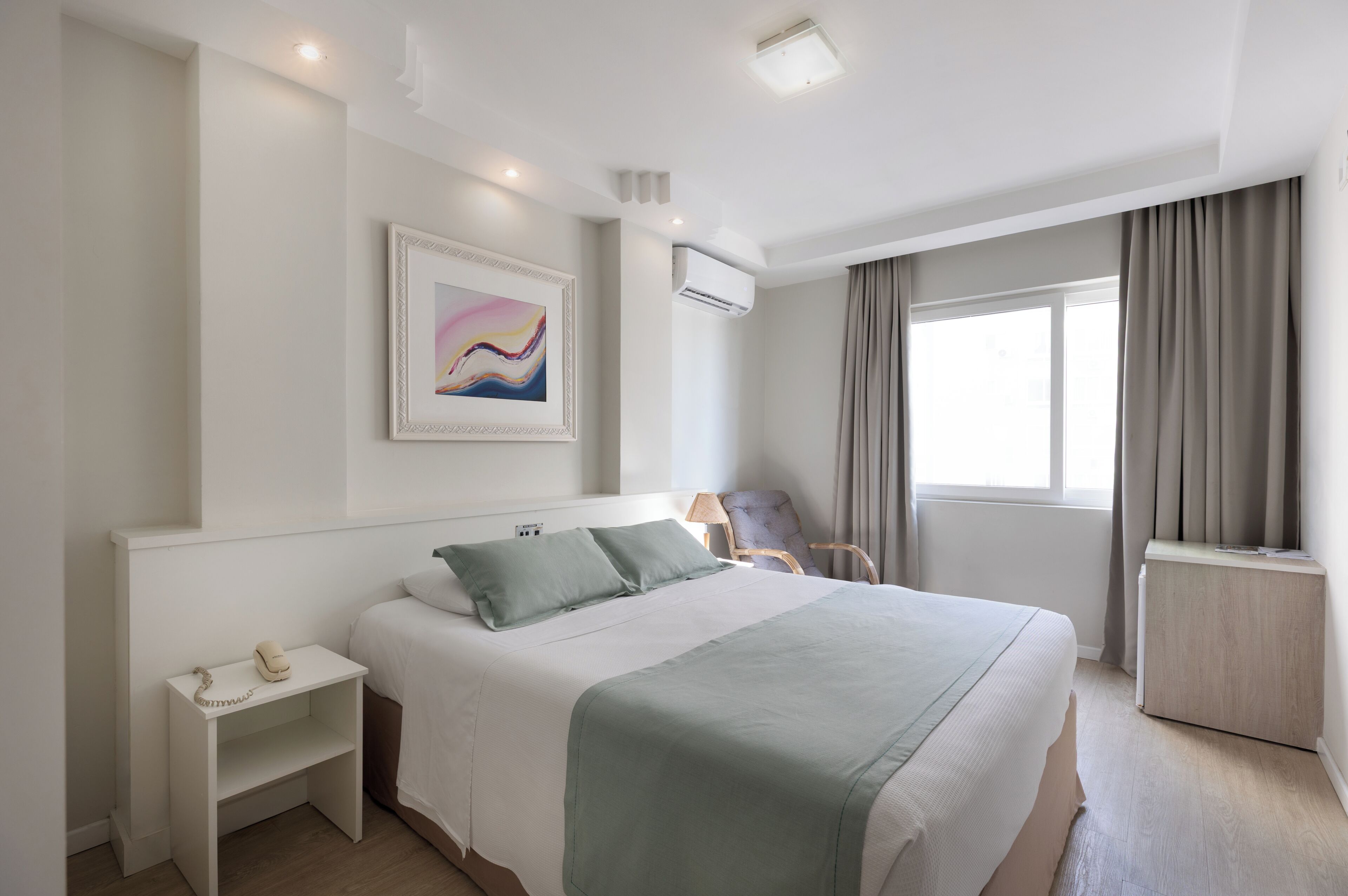 A hotel room with a bed, a bedside table with a telephone, a chair, a window with curtains, and an air conditioning unit.