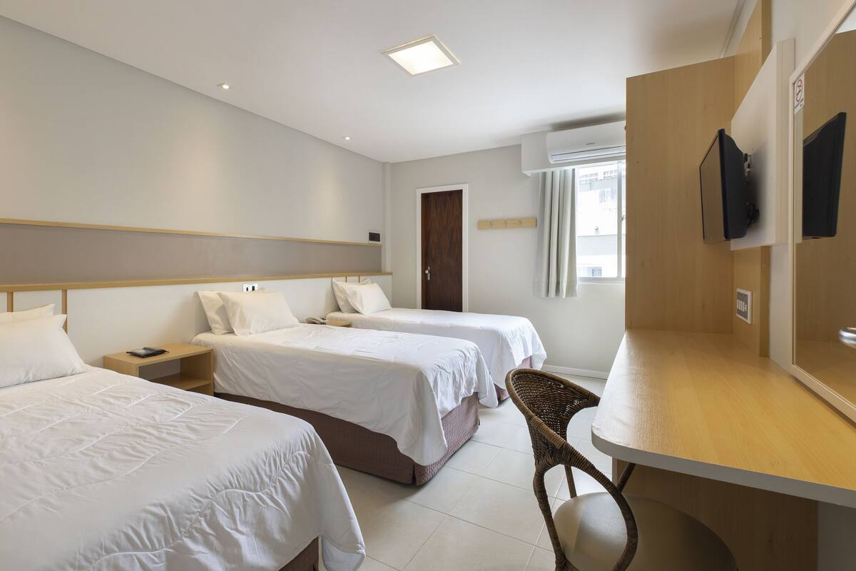 A hotel room with two beds, a wooden desk, a chair, a TV, and a window with curtains.