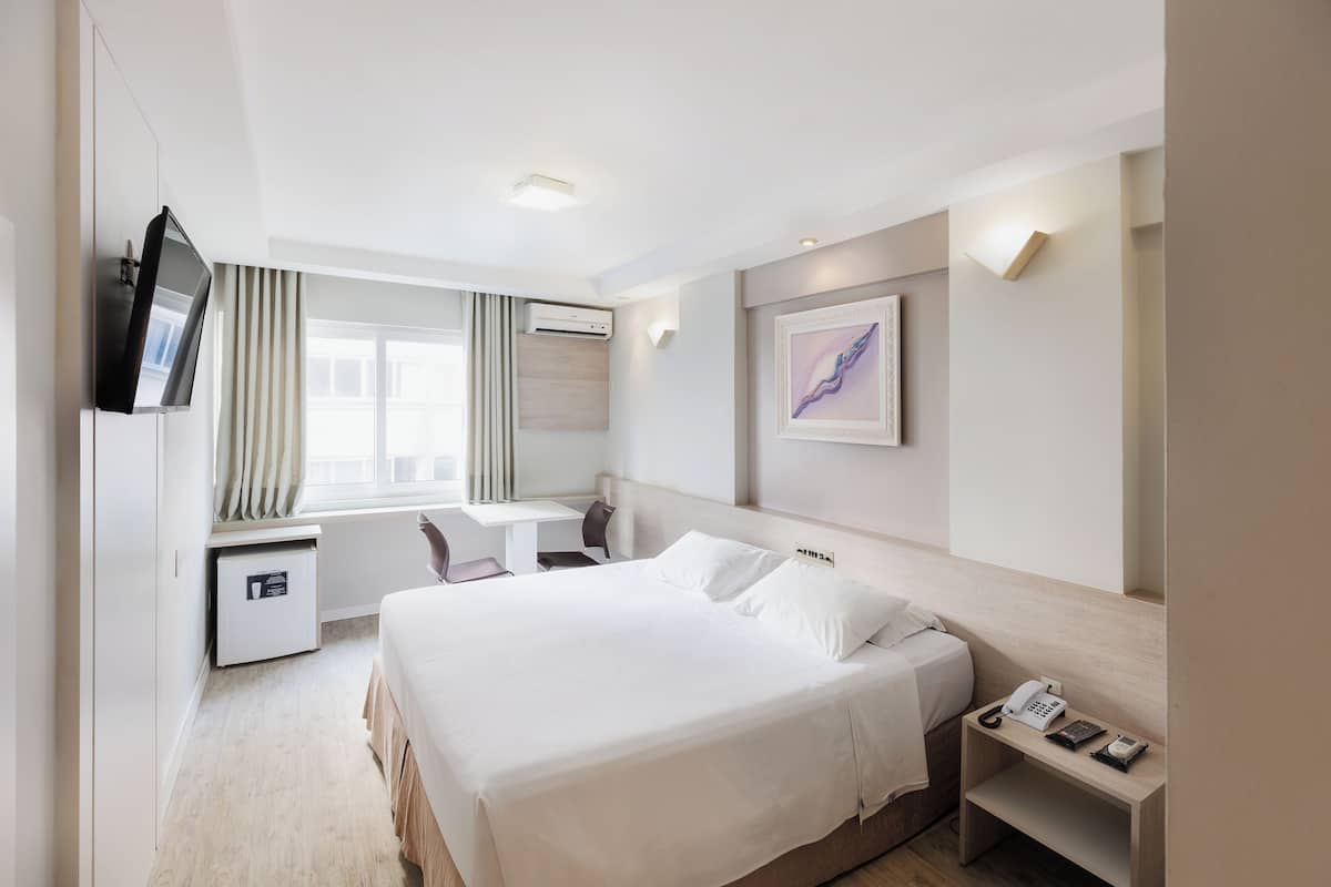 A hotel room with a bed, desk, chair, TV, and artwork on the wall.