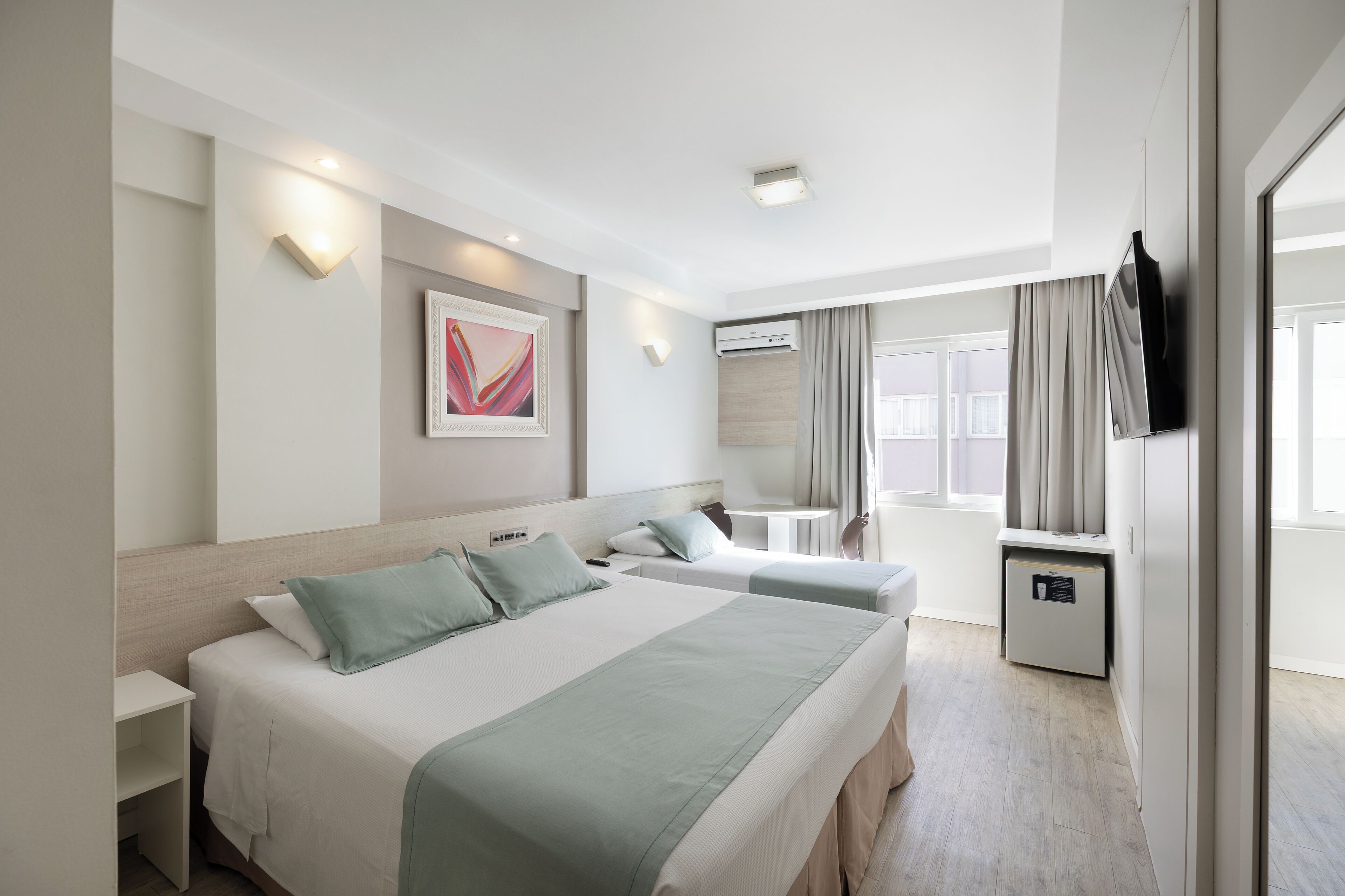 Habitación de hotel con dos camas, un escritorio, una silla, una mesita, un armario y una ventana con cortinas.
