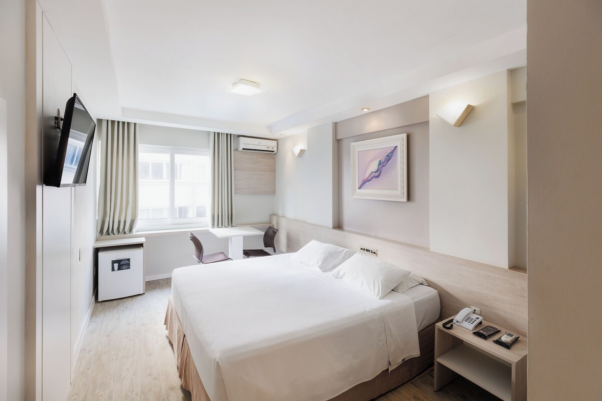 A hotel room with a bed, desk, chair, TV, and artwork on the wall.