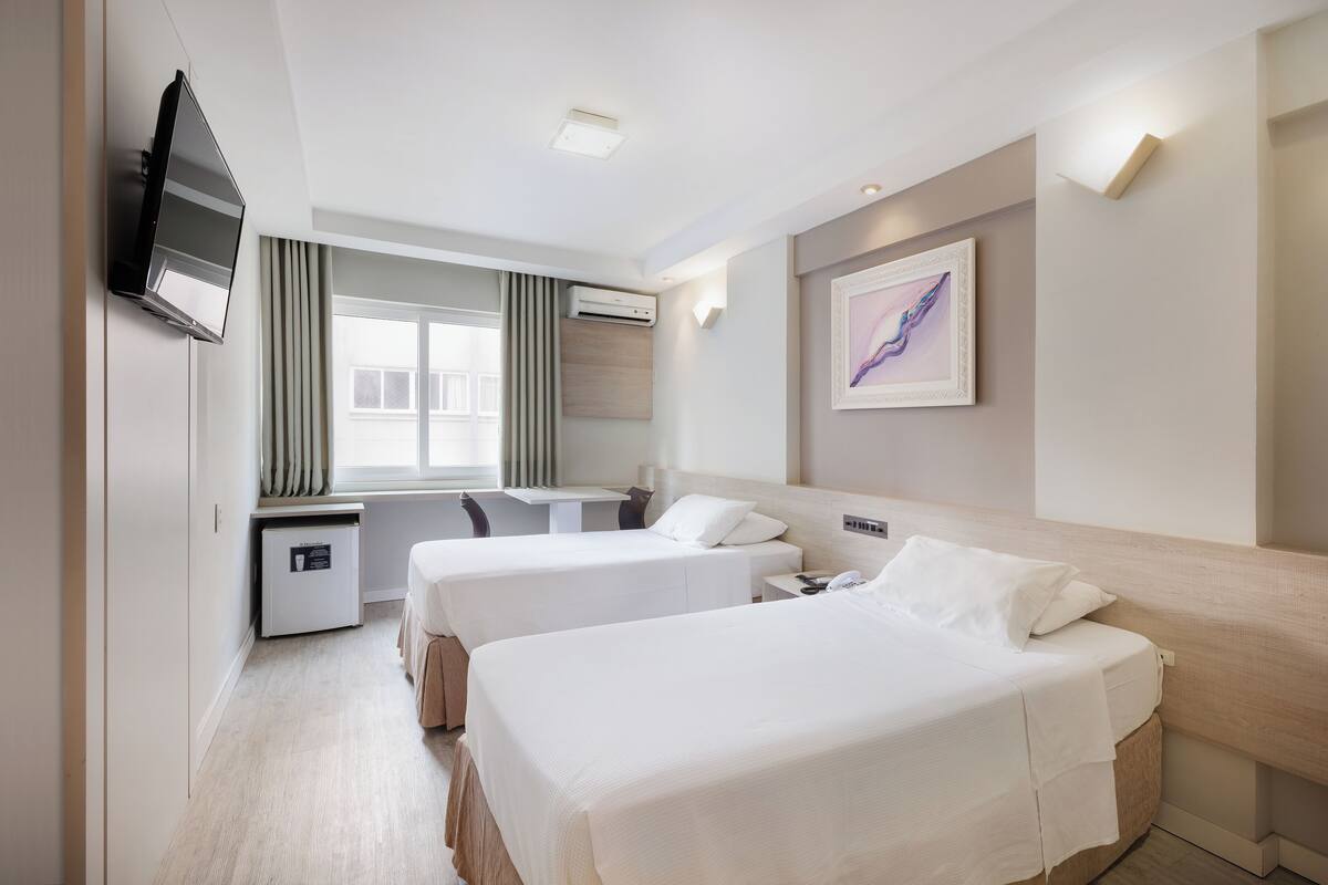 A hotel room with two beds, a desk, a TV, and a window with curtains.