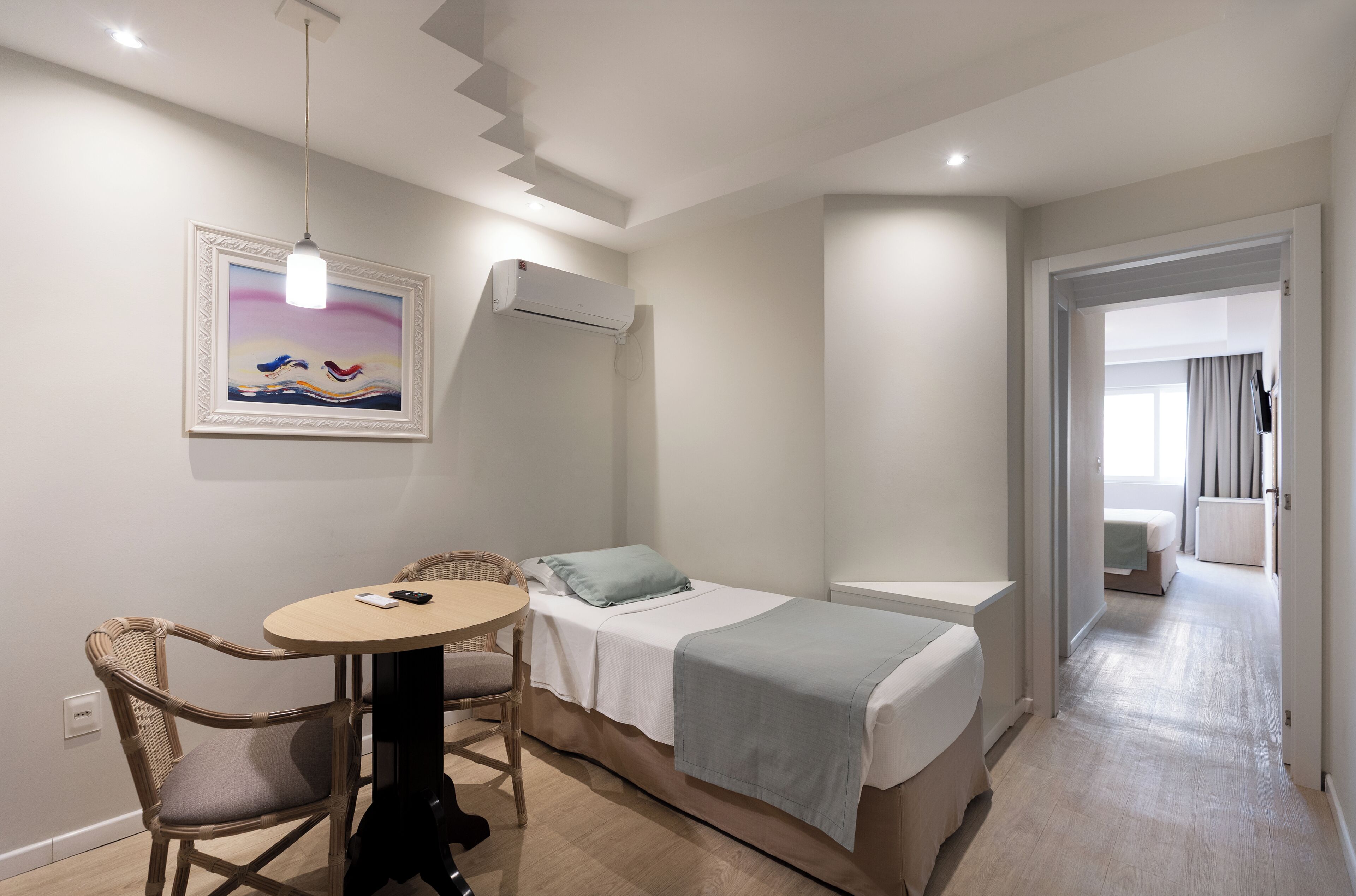 A hotel room with a bed, a small table, a chair, an air conditioning unit, a painting on the wall, and a view of the hallway.