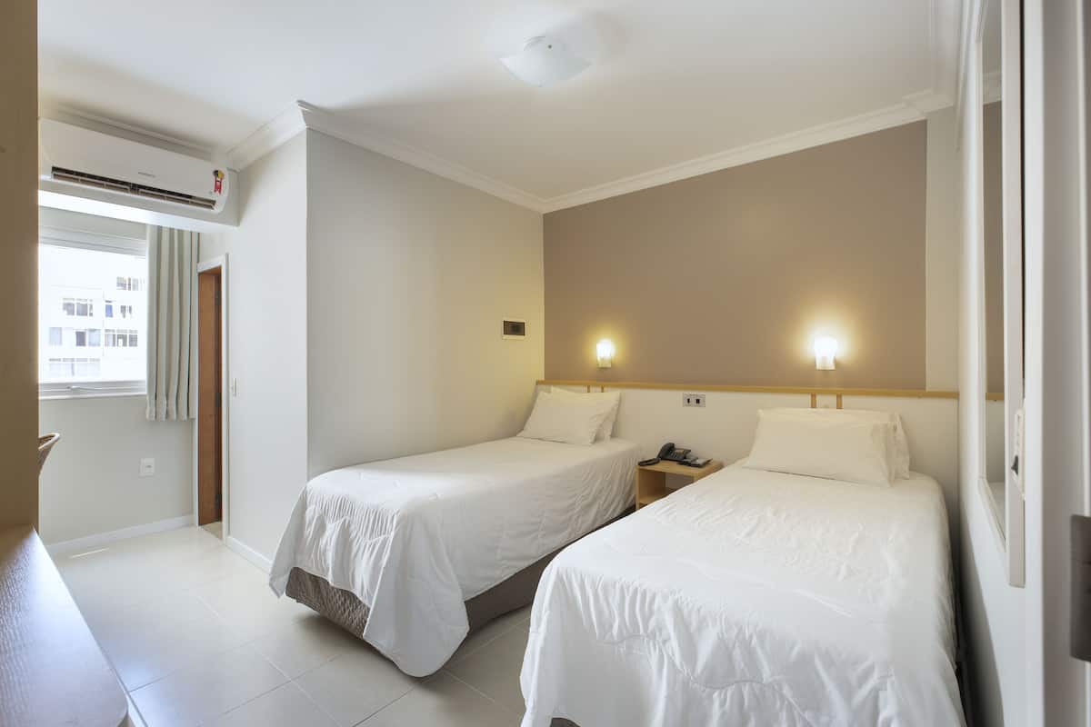 A hotel room with two single beds, a bedside table with a phone, a wall-mounted light, and an air conditioning unit.