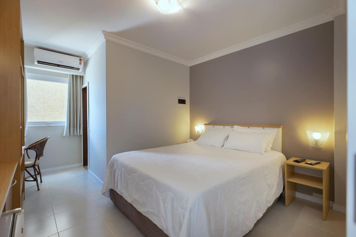 A hotel room with a bed, bedside table, chair, and an air conditioning unit.