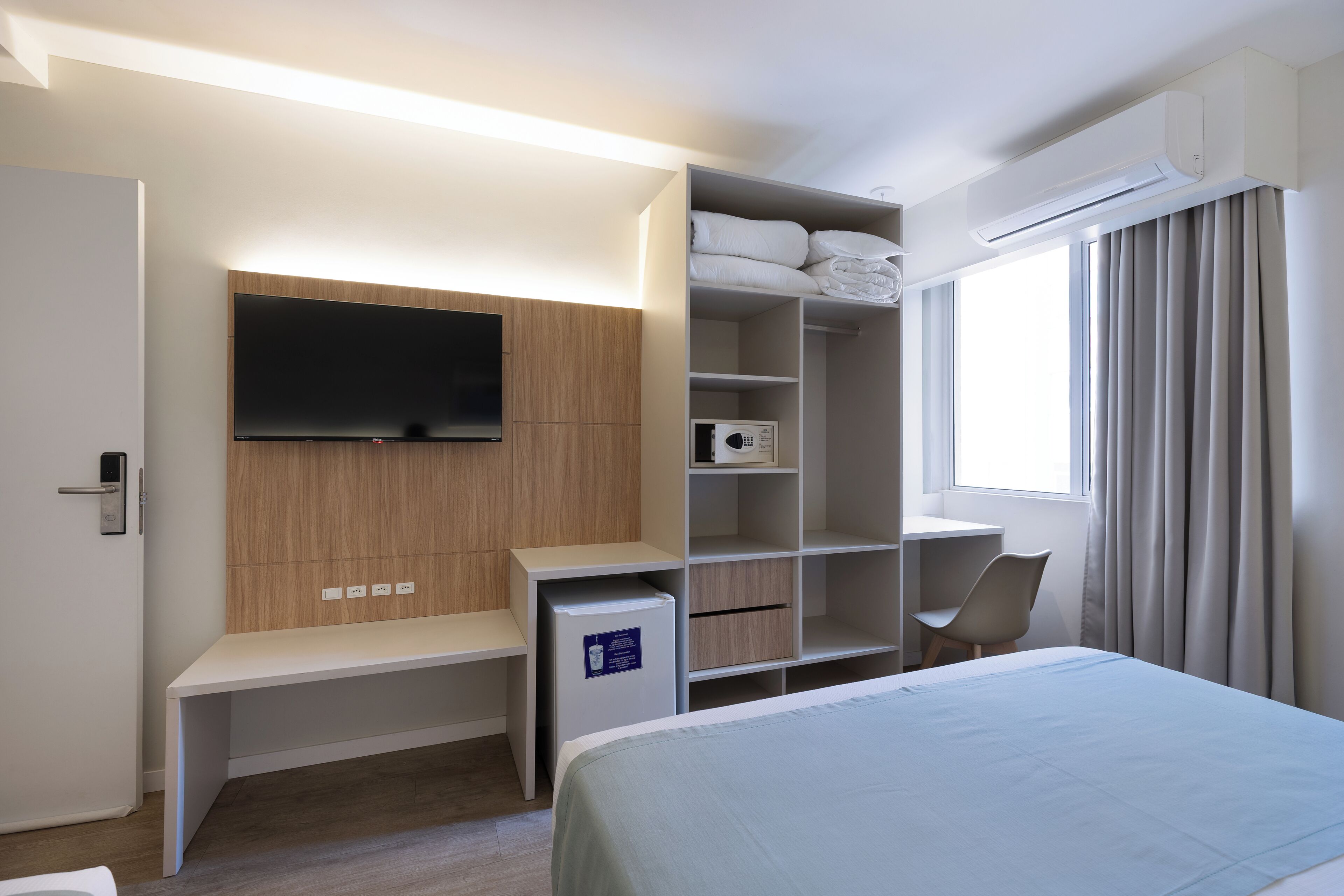 Een moderne hotelkamer met een bed, een televisie bevestigd aan een houten paneel, een bureau met een stoel, een minibar en een raam met gordijnen.