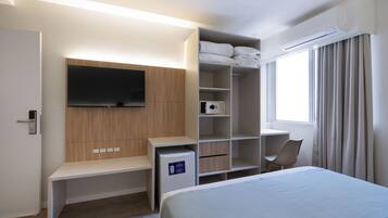 Een moderne hotelkamer met een bed, een televisie bevestigd aan een houten paneel, een bureau met een stoel, een minibar en een raam met gordijnen.