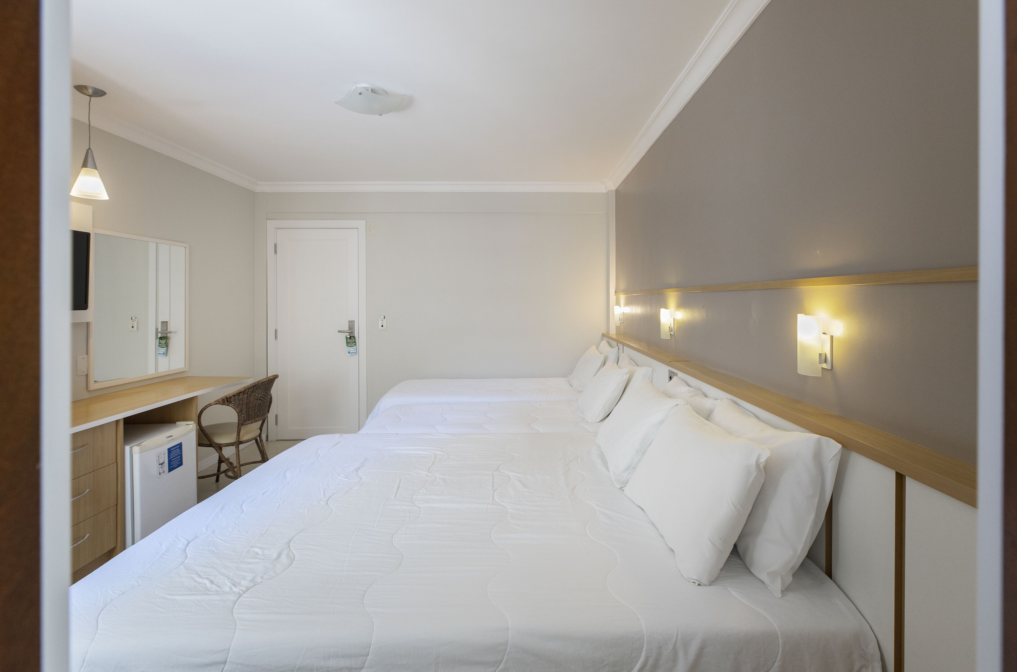 A hotel room with a large bed, a desk with a chair, a small refrigerator, and a mirror.