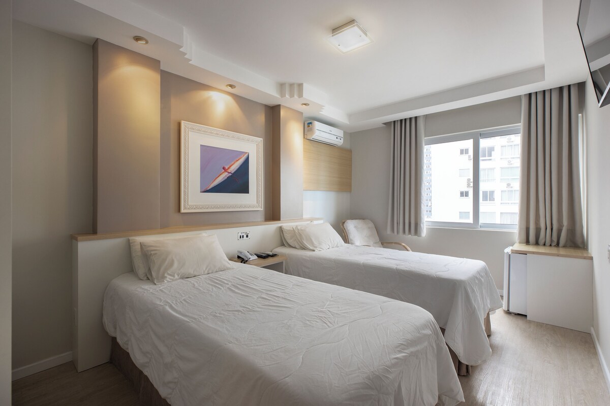 A hotel room with two beds, a bedside table, a wall-mounted air conditioner, a framed picture, and a window with curtains.