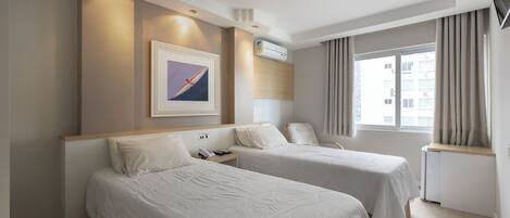 A hotel room with two beds, a mounted air conditioner, a framed picture on the wall, and a window with curtains.
