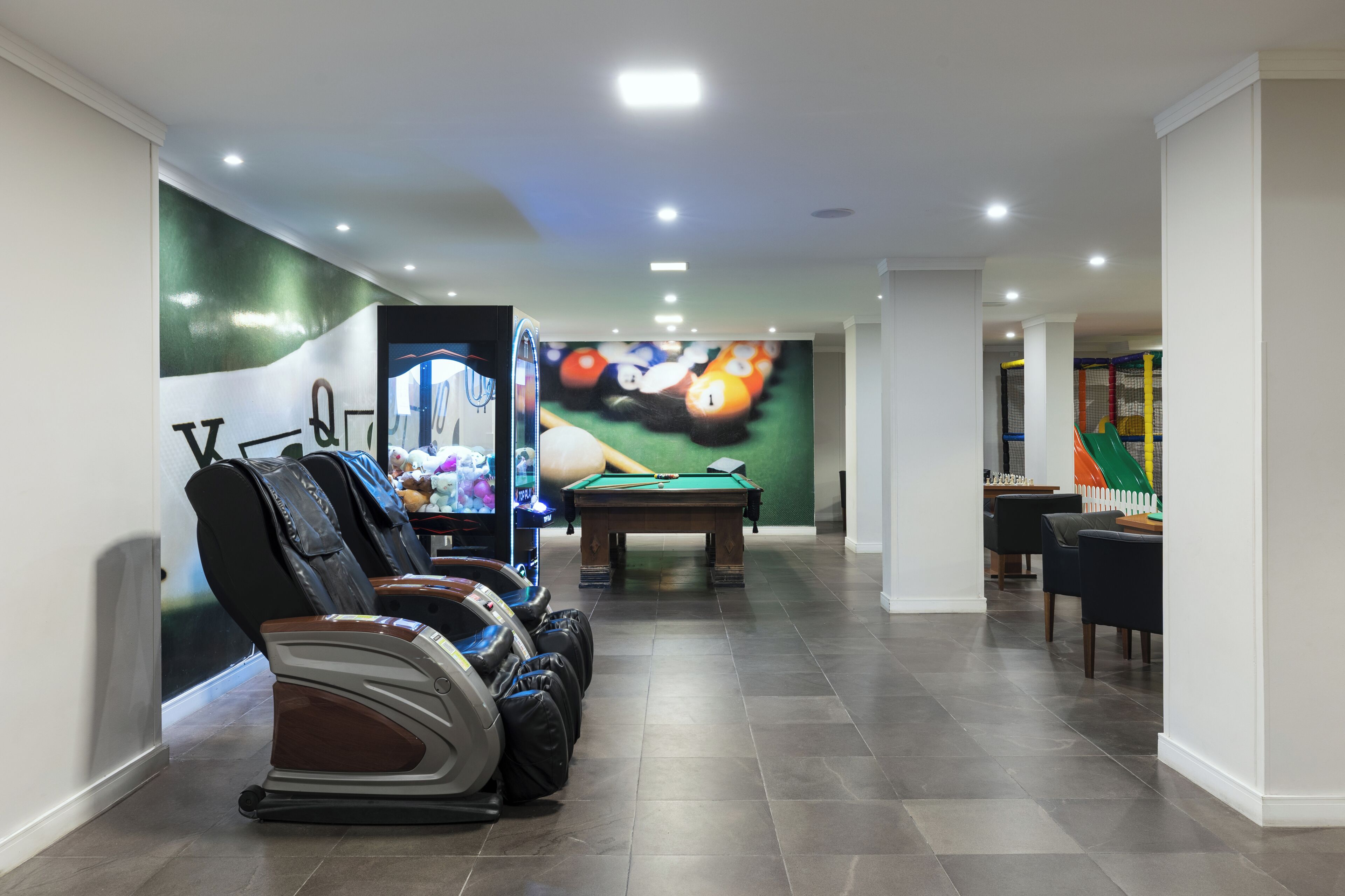 Mesa de bilhar em um espaço com cadeiras de massagem e um mural com tema de jogos de bilhar.
