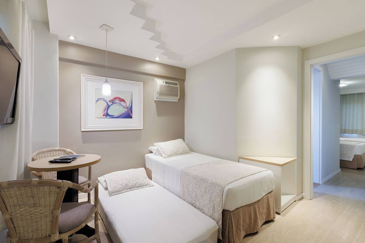 A hotel room with two single beds, a small round table with a chair, a wall-mounted air conditioner, and a framed abstract painting.