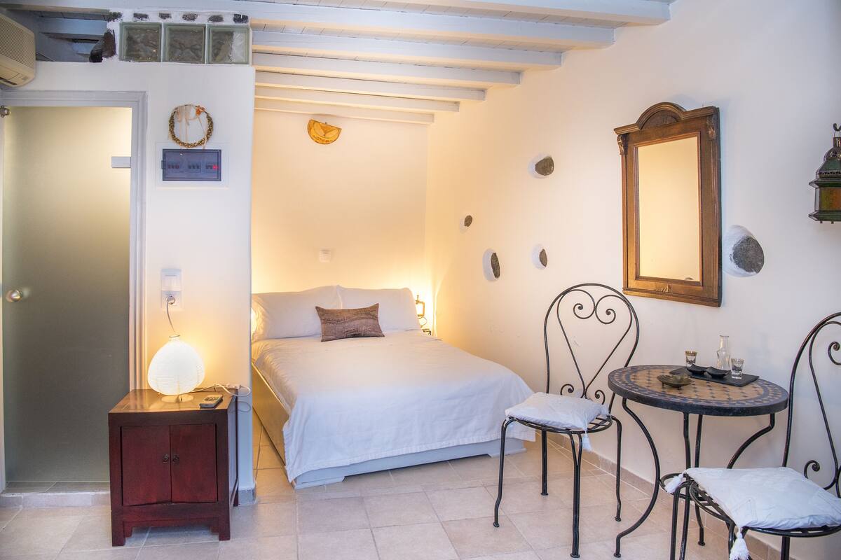 Een slaapkamer met een bed, nachtkastje, lamp en een klein rond tafeltje met stoelen.