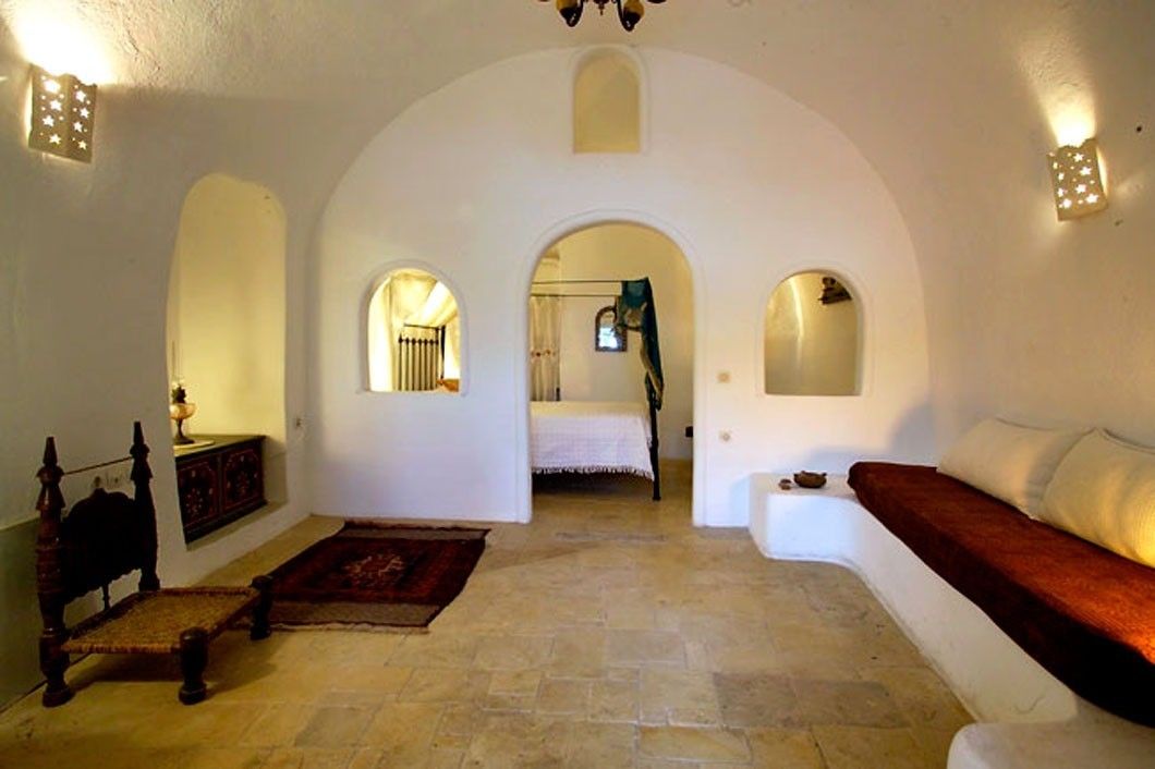 A room with a bed, a wooden chair, a sofa, and a small table. There are two arched doorways leading to another room.