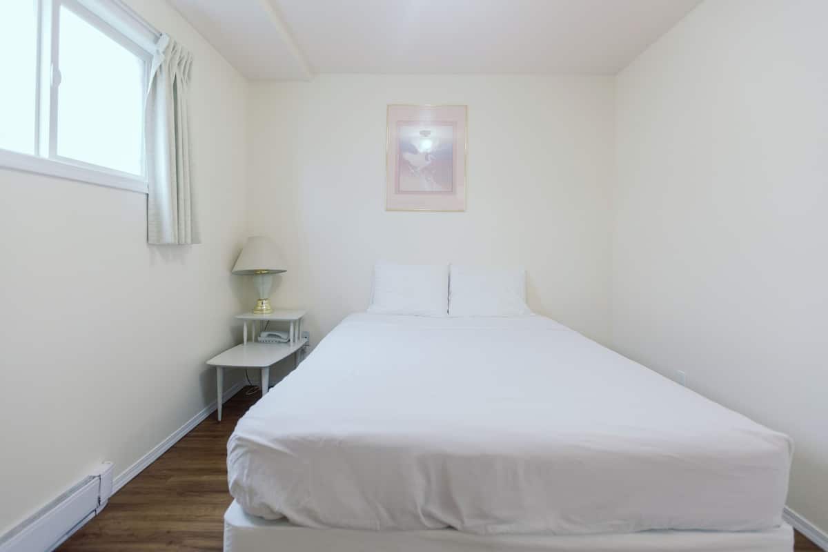 Une petite chambre propre, avec un lit simple, une table de chevet avec une lampe et une fenêtre avec des rideaux.