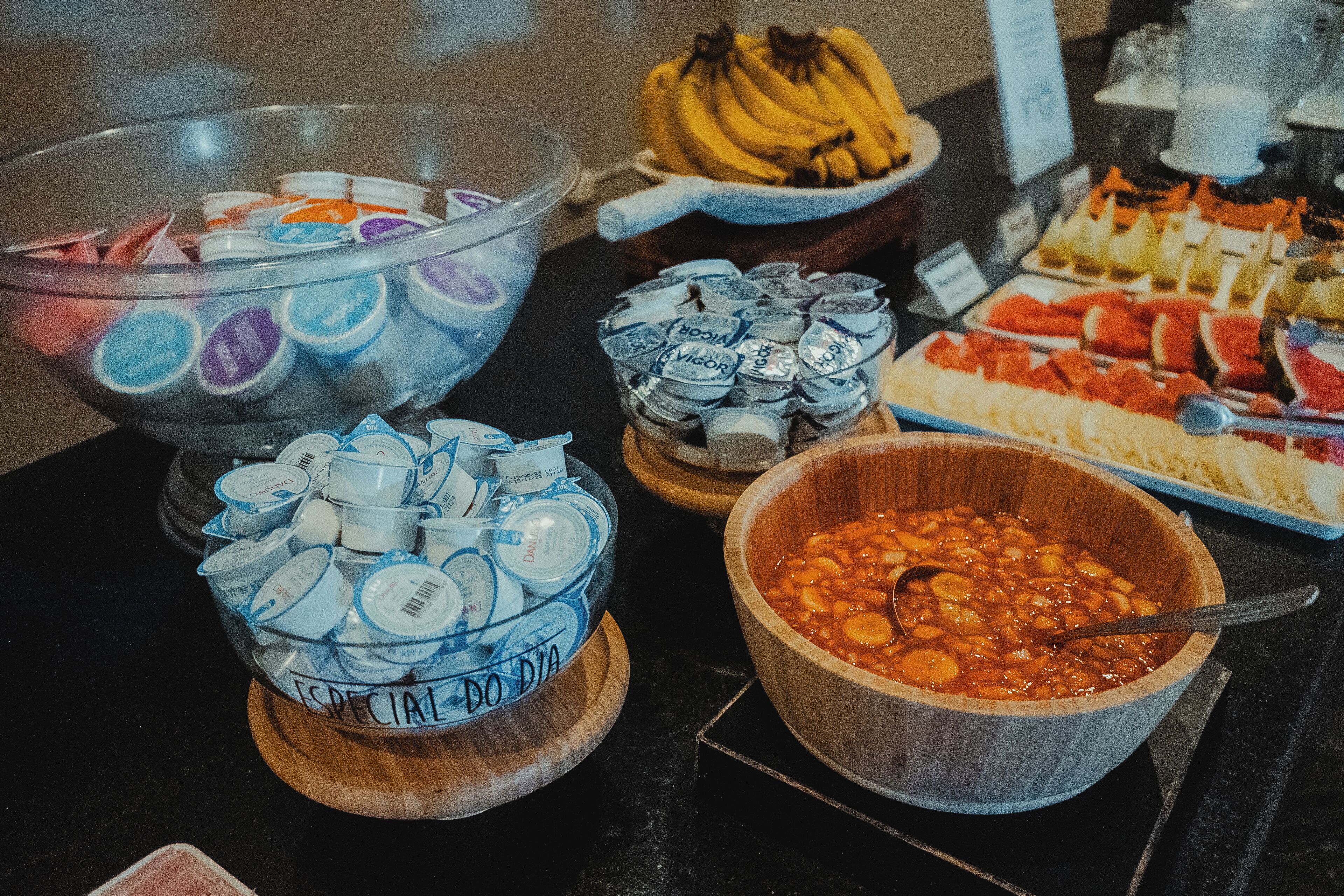 Un bufé con diversos alimentos, incluyendo un tazón de frijoles, un tazón de yogur, un tazón de helado, una bandeja de frutas y un tazón de plátanos.