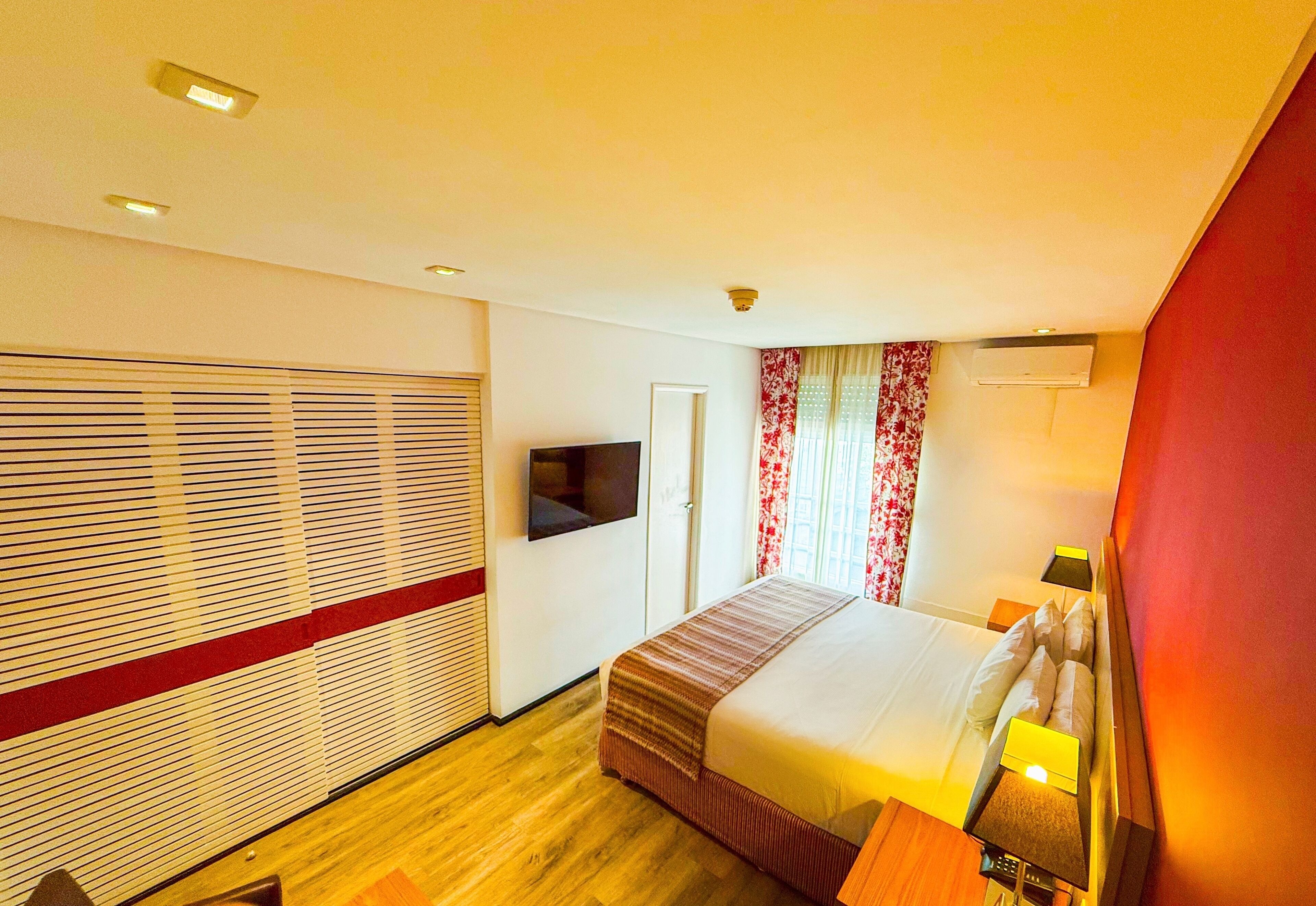Quarto de hotel com cama, televisão presa na parede, janela com cortinas e piso de madeira.