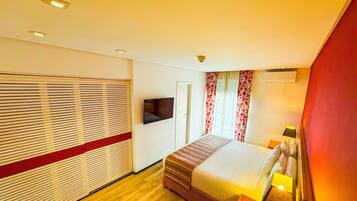 Quarto de hotel com cama, televisão presa na parede, janela com cortinas e piso de madeira.